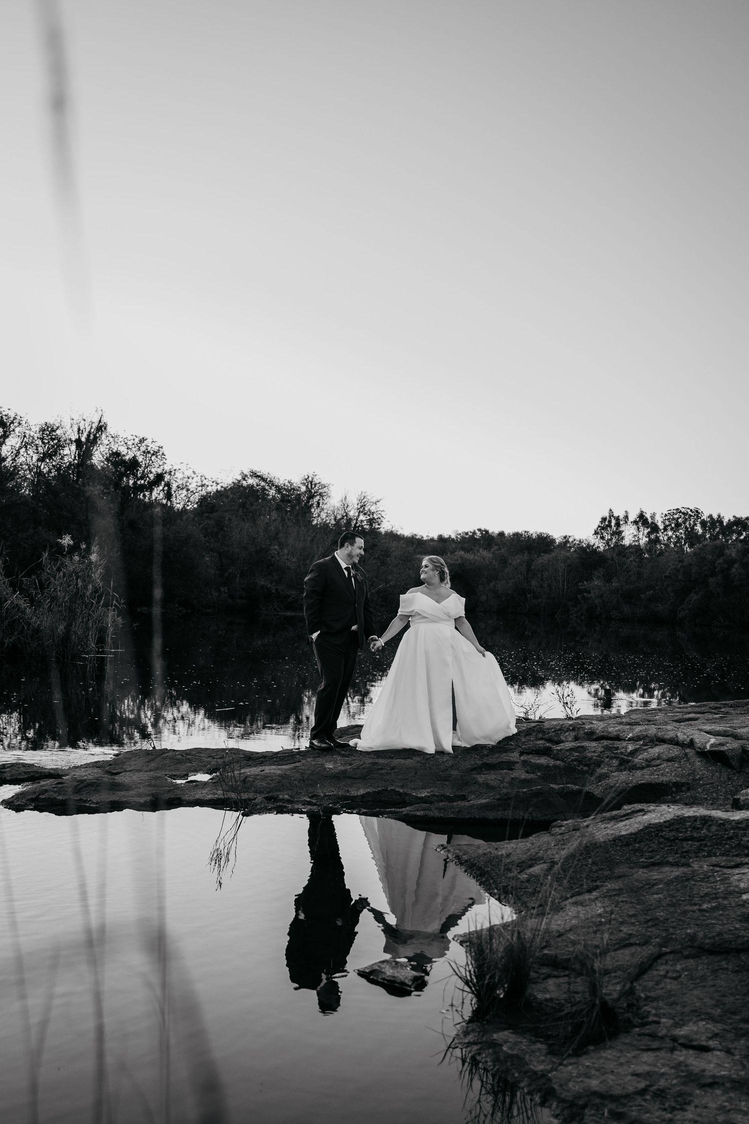 Black and white wedding photo of bride and groom holding hands by a reflective pond with trees in background.