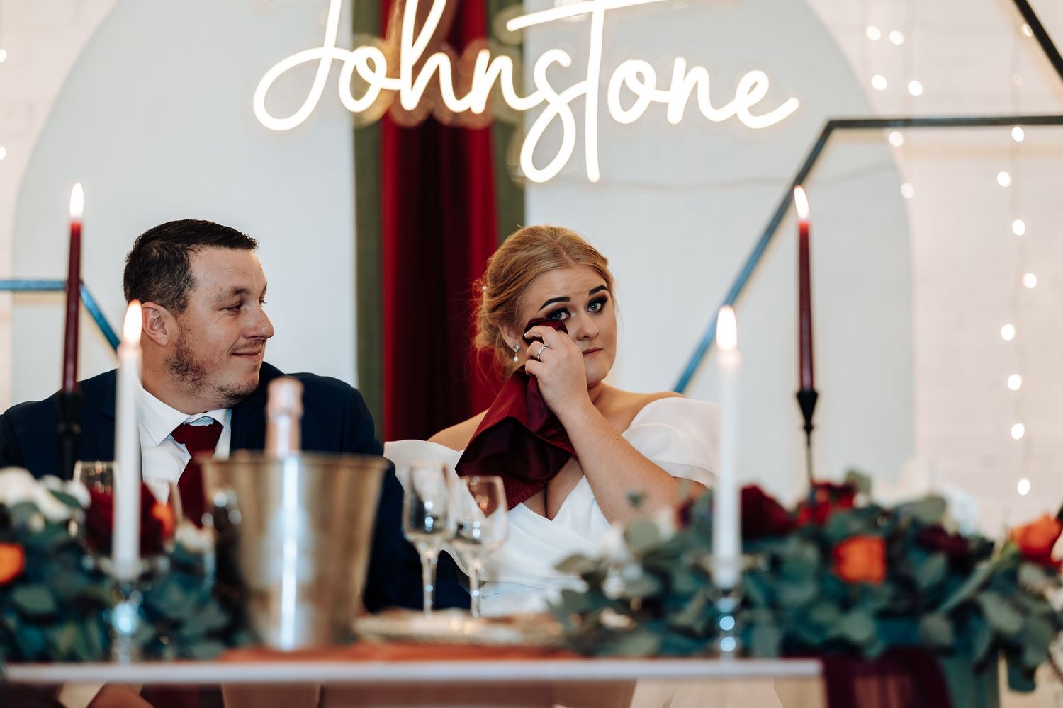 Bride and groom at wedding reception table beneath neon Johnstone sign, surrounded by candles and floral arrangements.