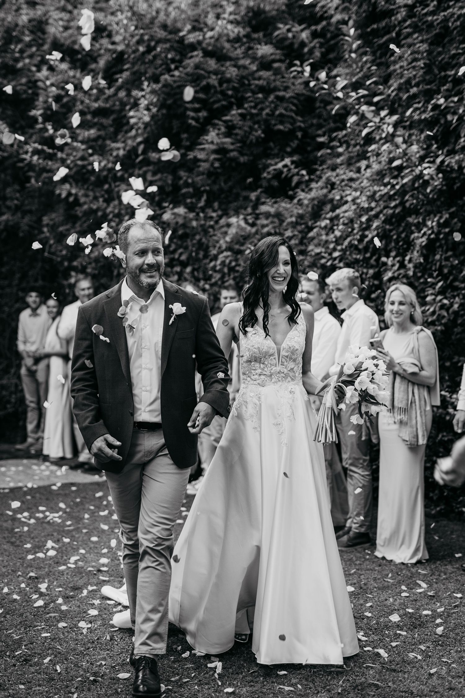 Black and white photo of a smiling bride and groom walking through confetti with wedding guests cheering behind them.