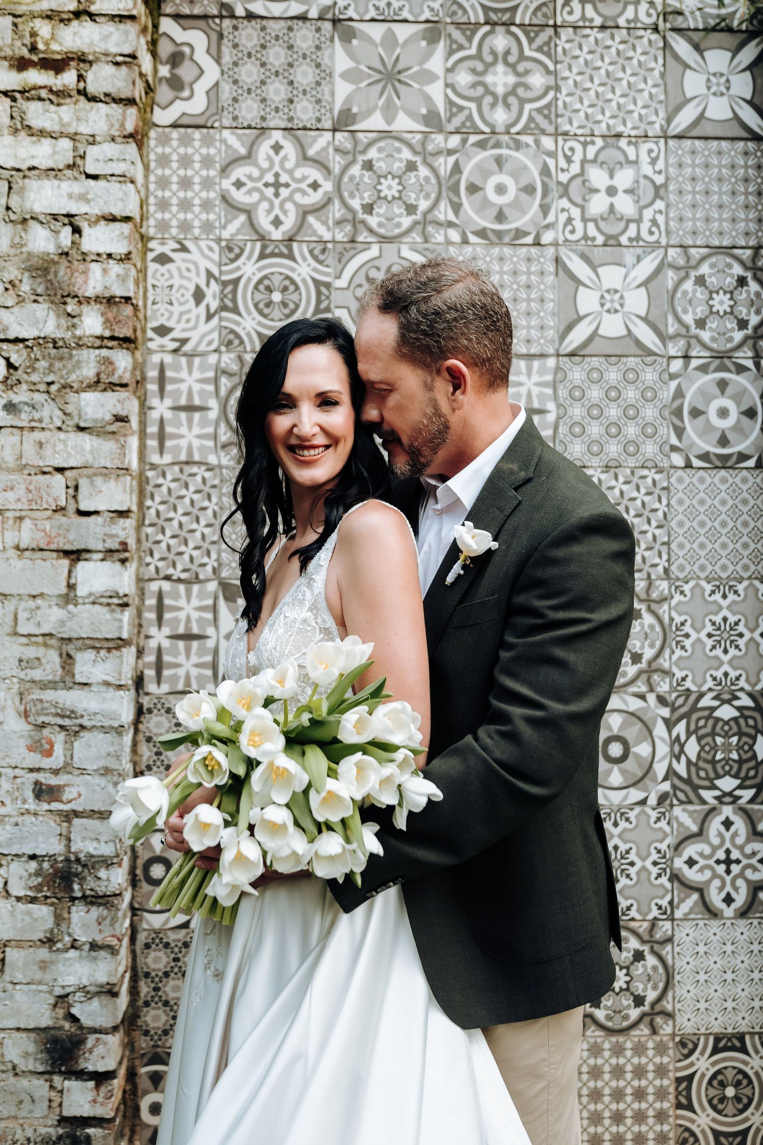 Bride and groom embrace before decorative tile wall, bride holding white tulip bouquet in elegant wedding attire.