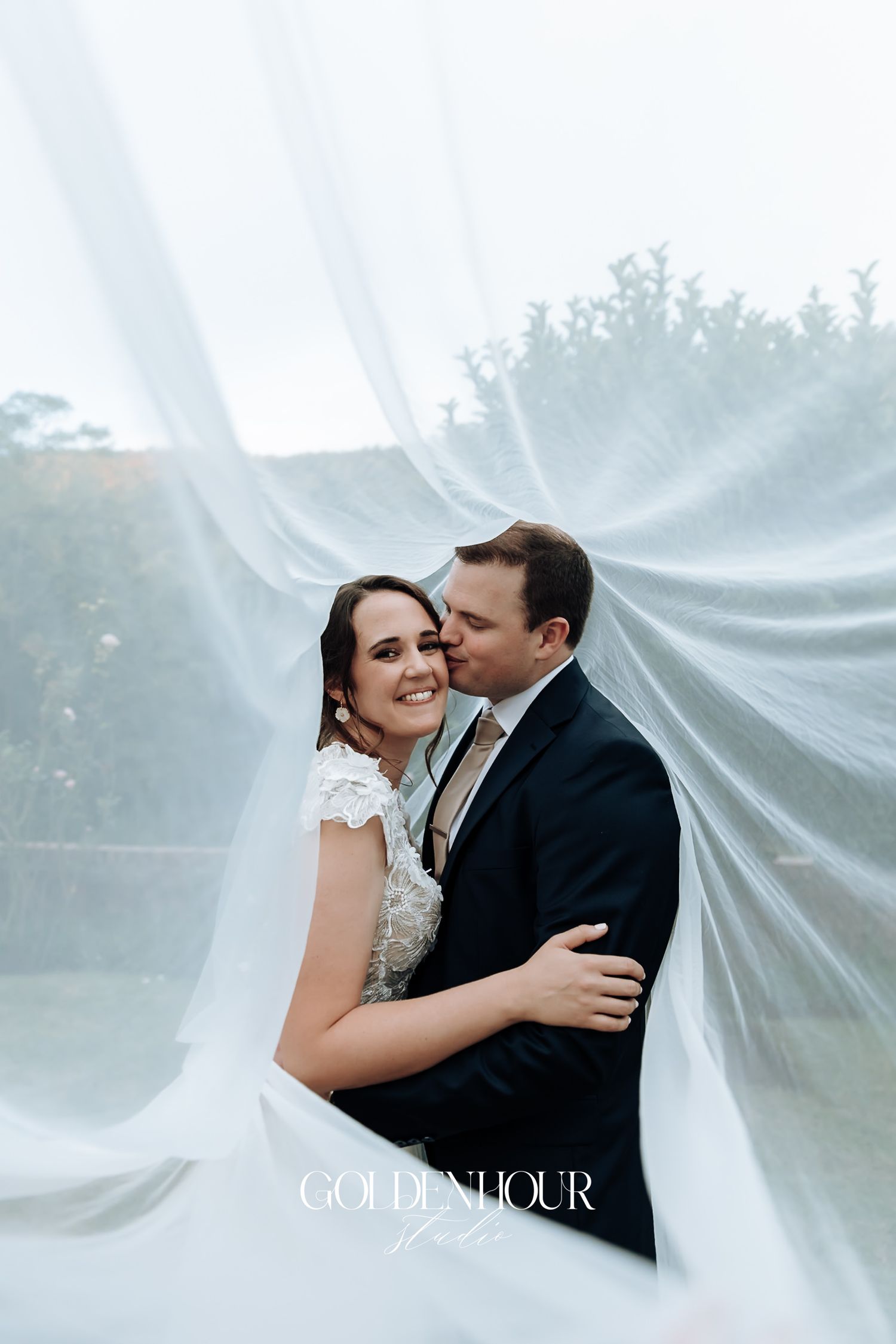 Groom embracing smiling bride wrapped in flowing white veil in dreamy outdoor wedding portrait.