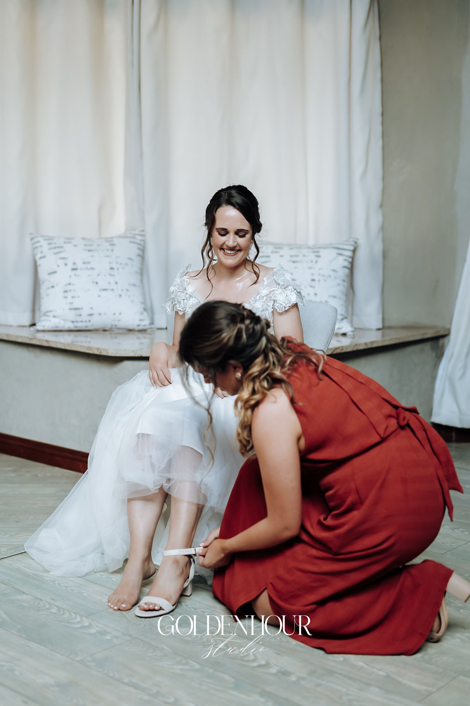 Bridesmaid in red dress helps bride with wedding shoes while bride sits smiling in white gown.