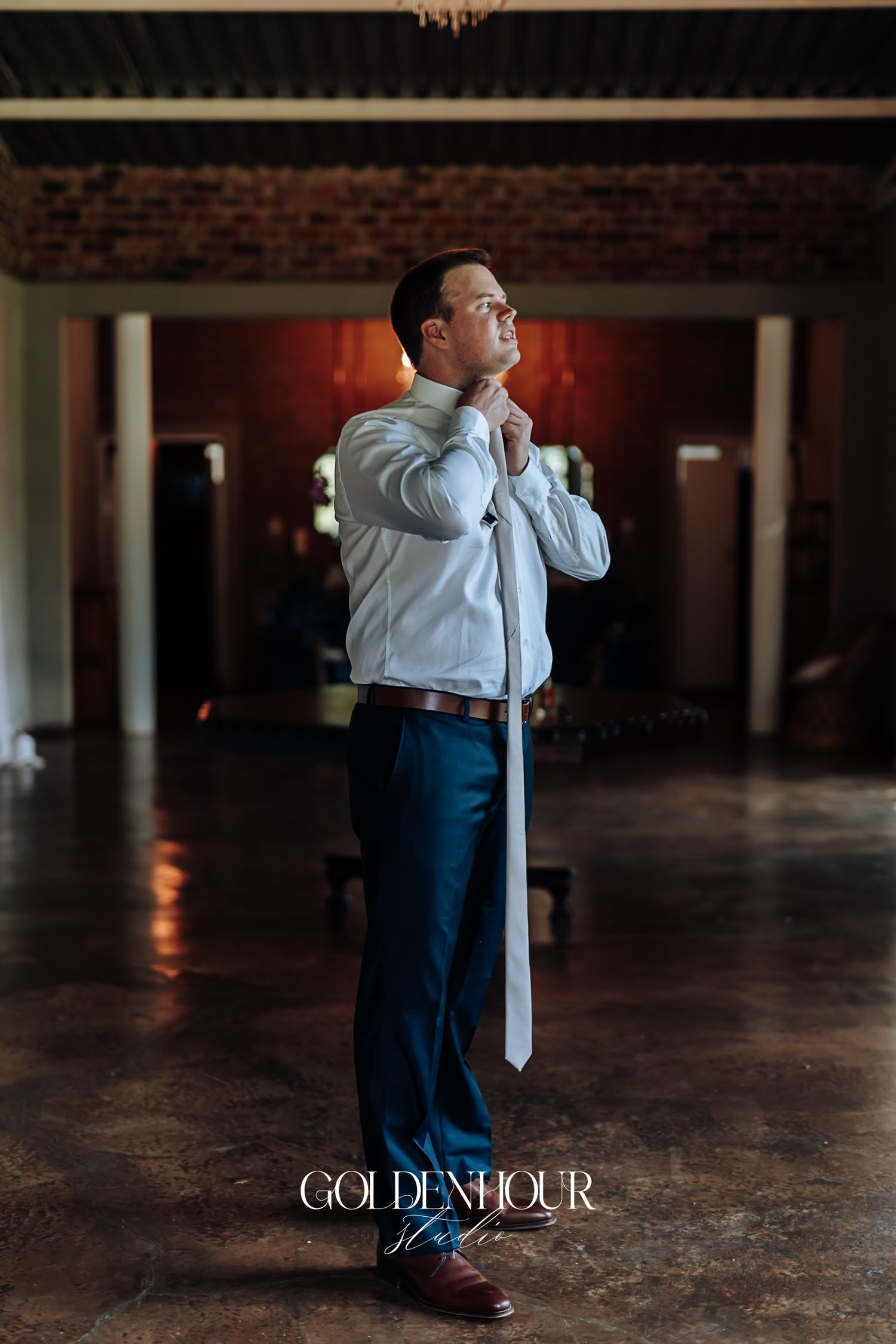 Groom adjusting his white tie while getting ready in a rustic brick-walled wedding venue.