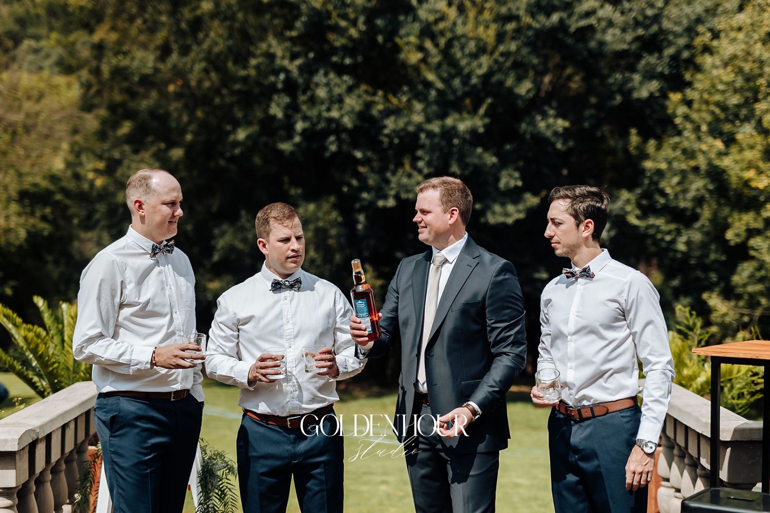 Four groomsmen in navy pants and white shirts socializing outdoors at a wedding.