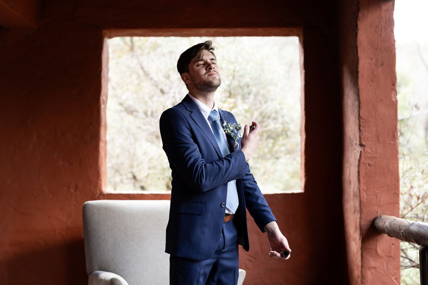 Man in navy suit adjusting boutonniere by window.