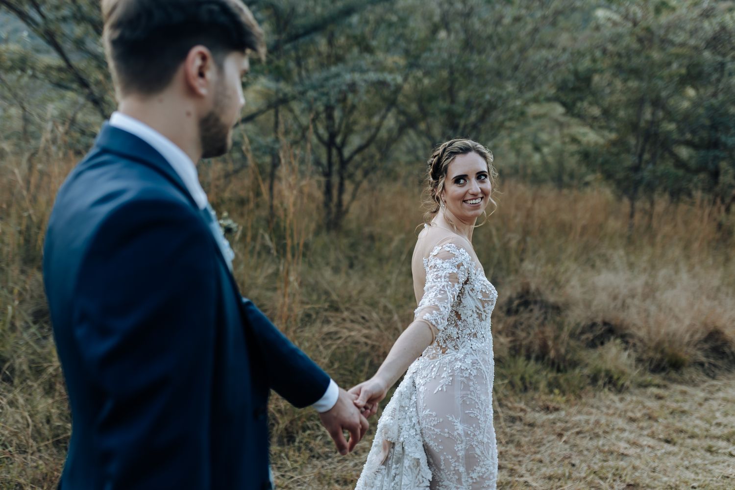 A romantic wedding couple holding hands in a rustic outdoor setting with moody lighting and natural scenery.