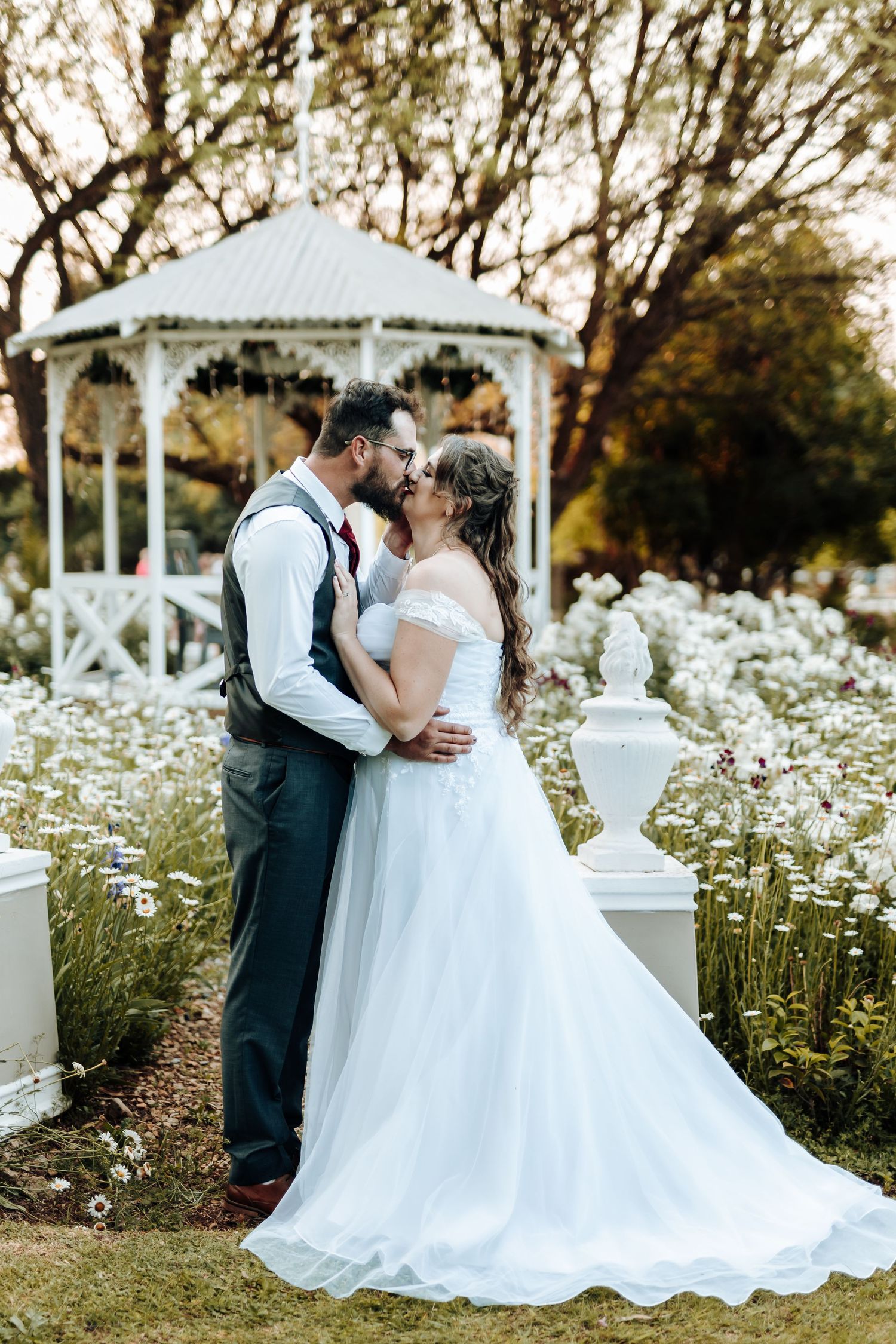 Romantic wedding couple embracing by white gazebo surrounded by wildflowers at sunset.