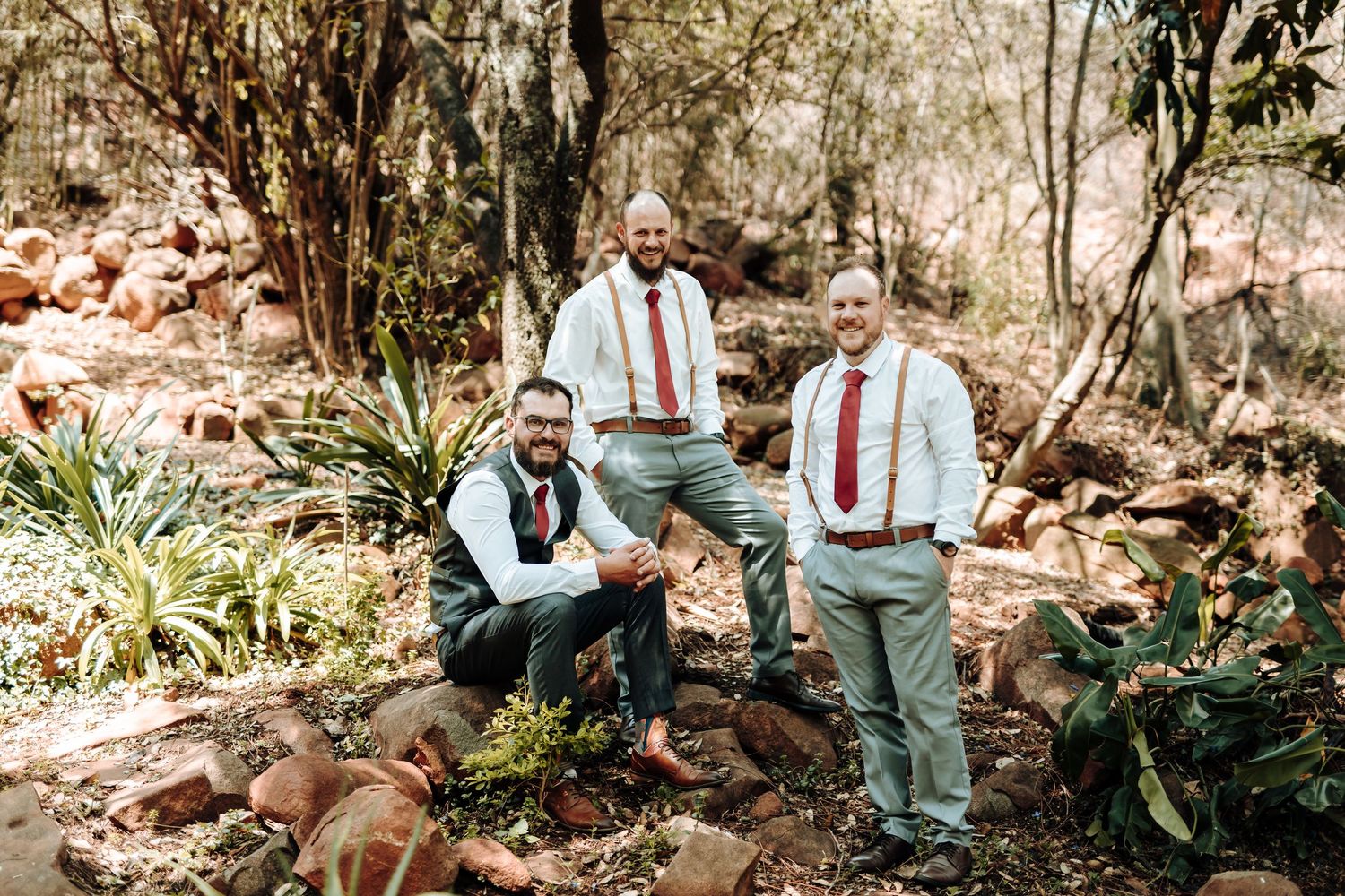 Three groomsmen in coordinating attire pose casually on rocky outdoor terrain.