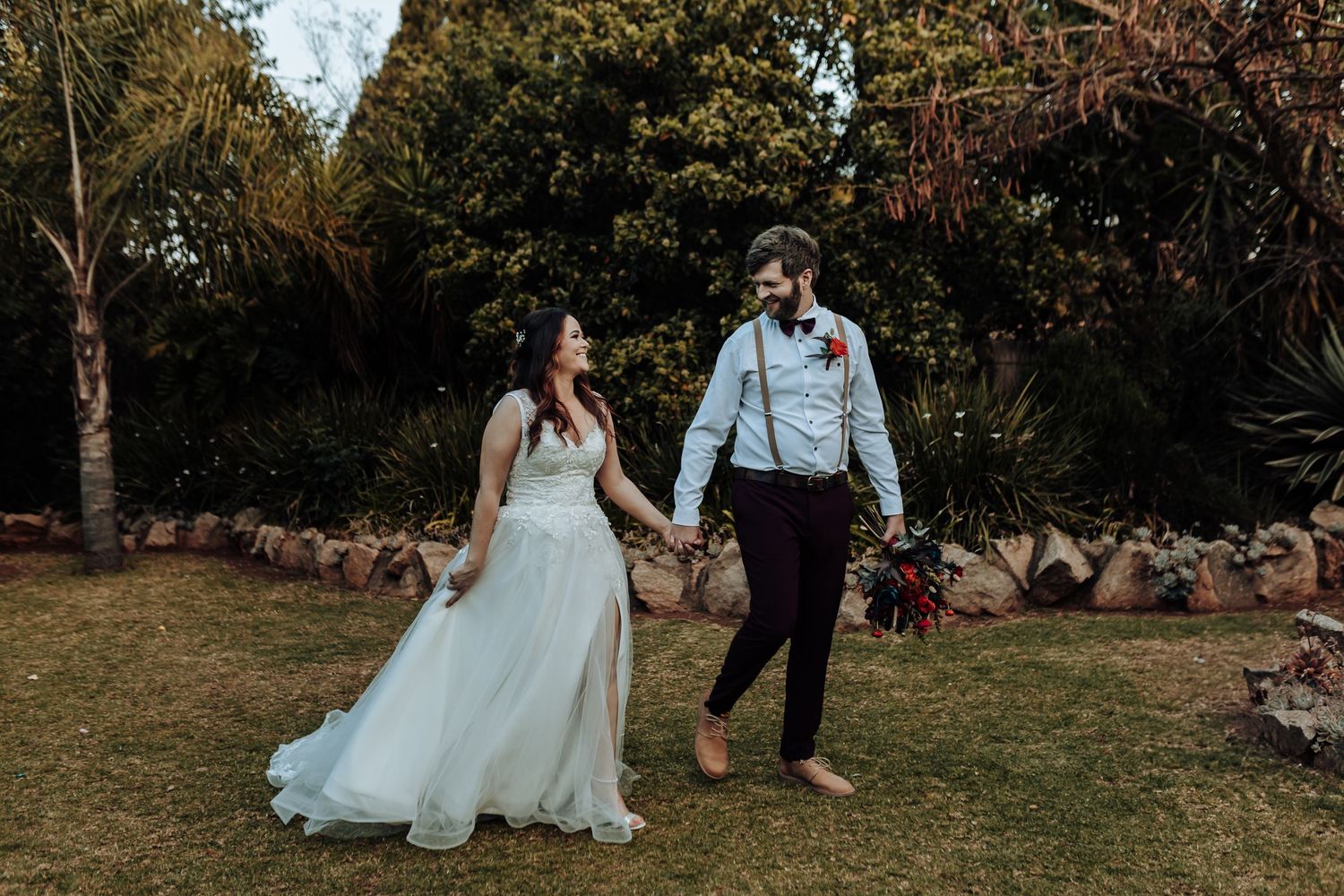 Couple walking hand in hand on grass path in wedding attire near stone wall.