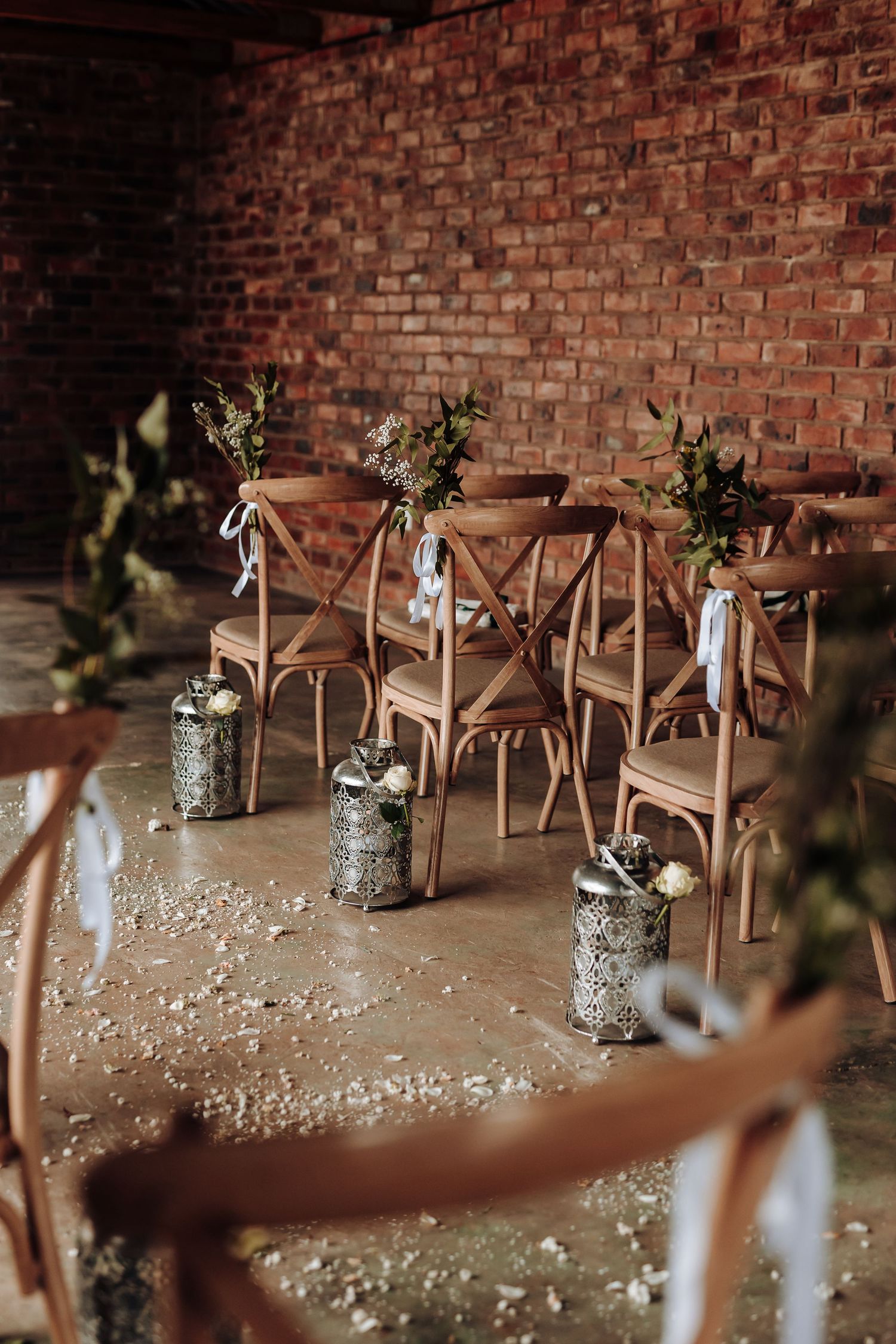 Close-up of vintage wooden chairs decorated with greenery and mason jars in a brick-walled venue.