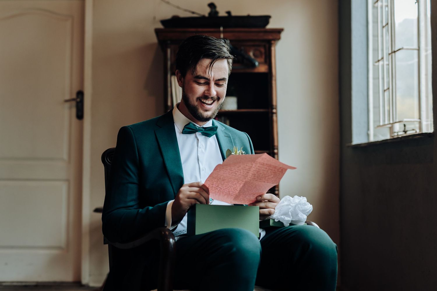 A person in a teal tuxedo and bowtie smiles while reading from a pink card in an elegant indoor setting.