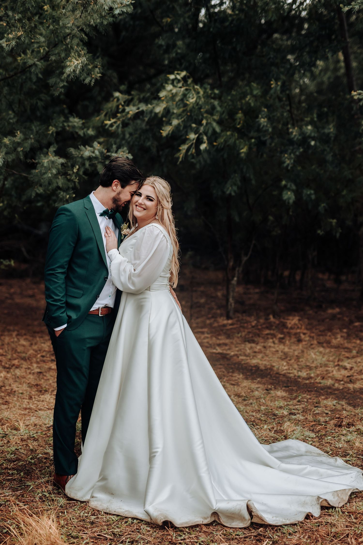Elegant wedding couple poses in forest setting with bride's dress trailing behind.