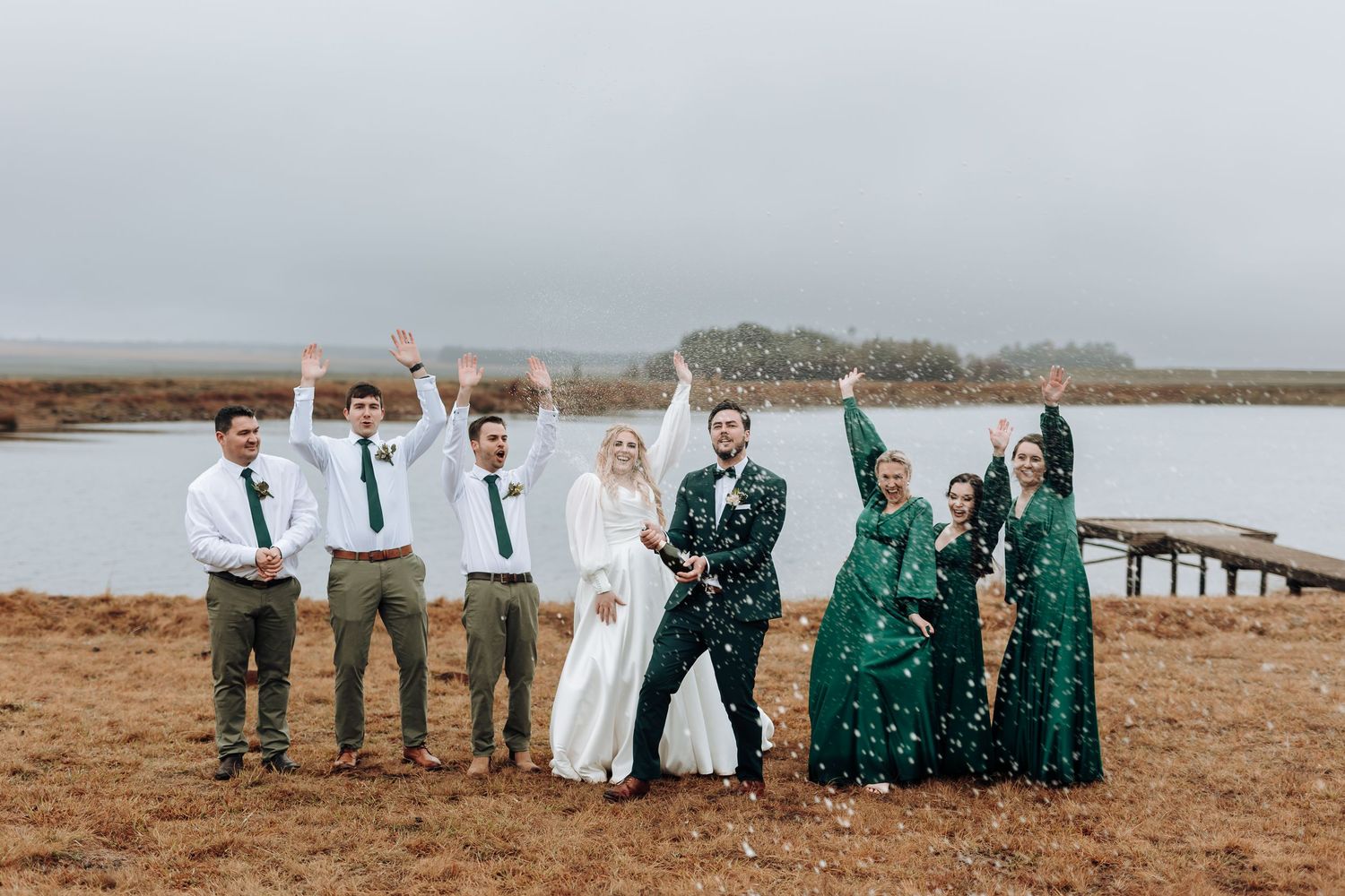 Wedding party poses playfully on a beach boardwalk with groomsmen in white shirts and bridesmaids in emerald dresses.