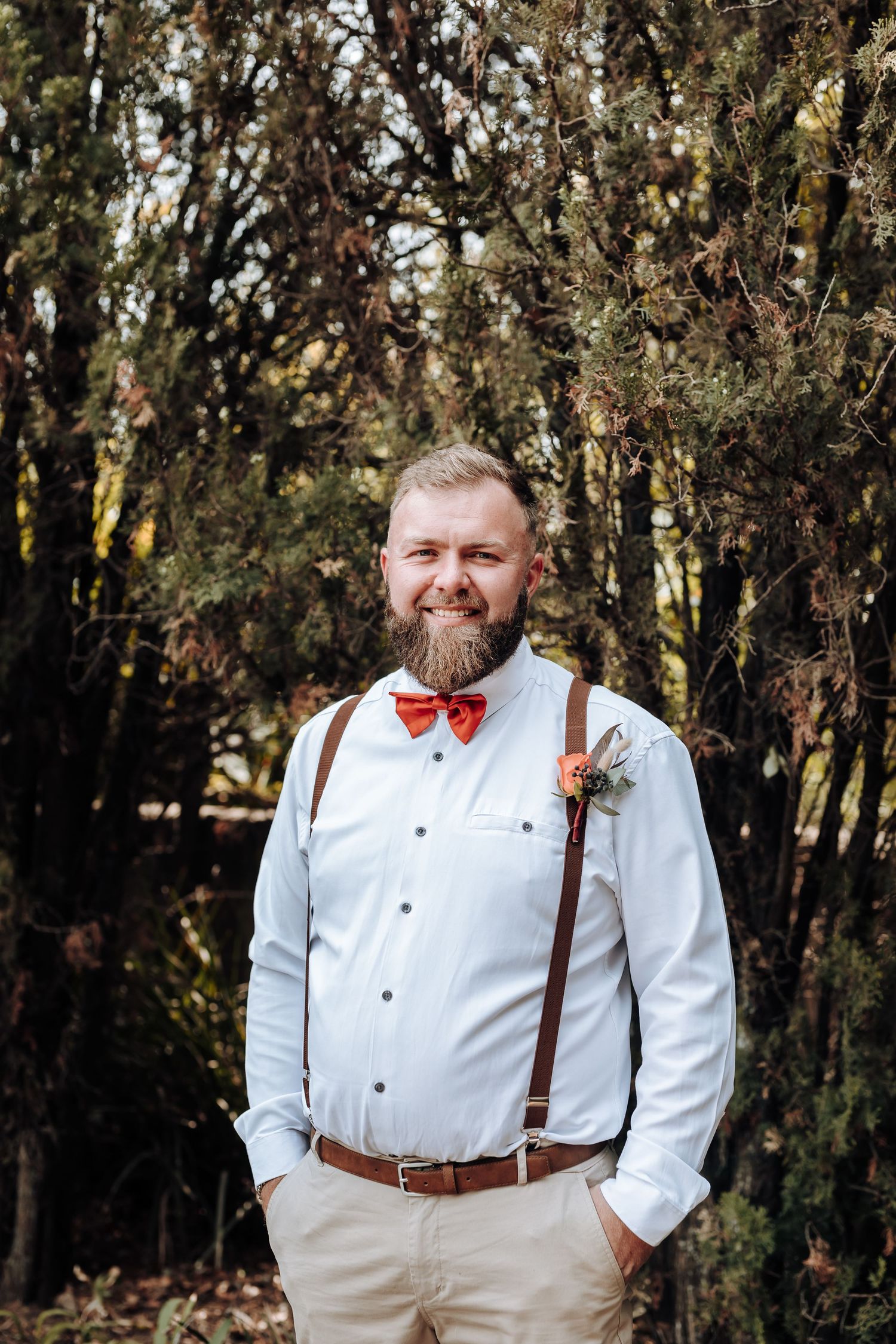 Groomsman in white shirt with suspenders and red bow tie smiles in natural setting.
