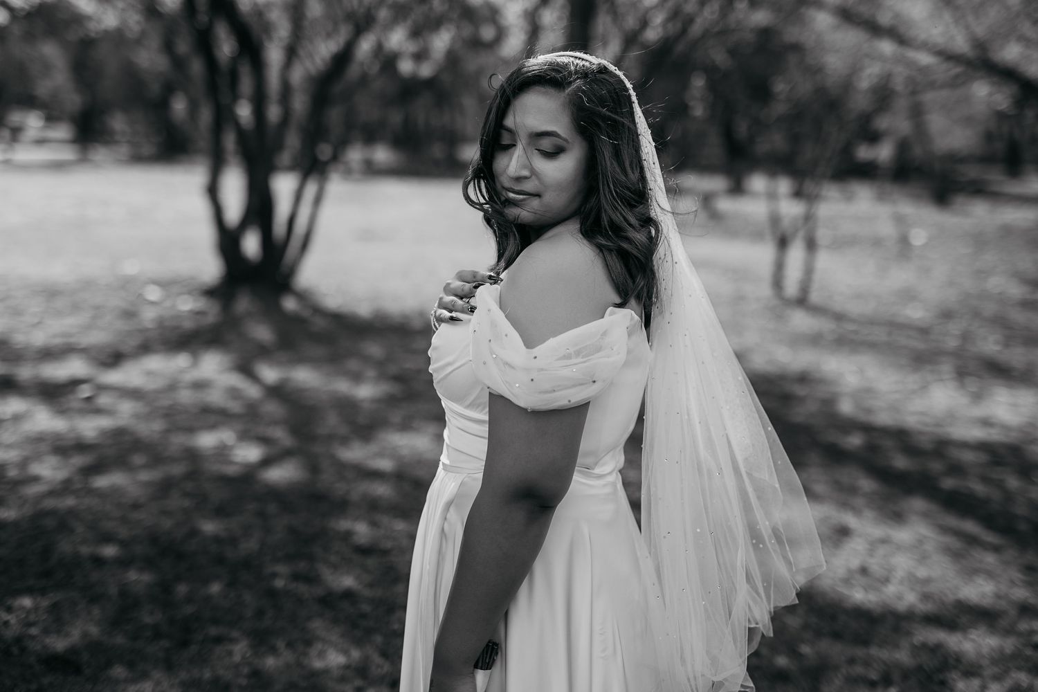 Bride in off-shoulder wedding dress poses in outdoor garden setting.