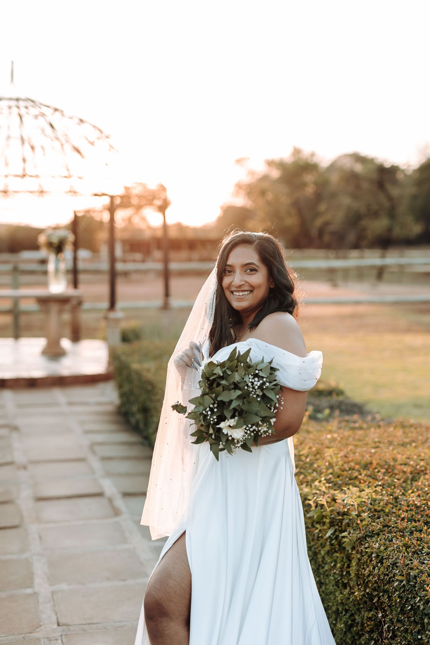 Radiant bride in off-shoulder gown holds eucalyptus bouquet during golden hour photoshoot.