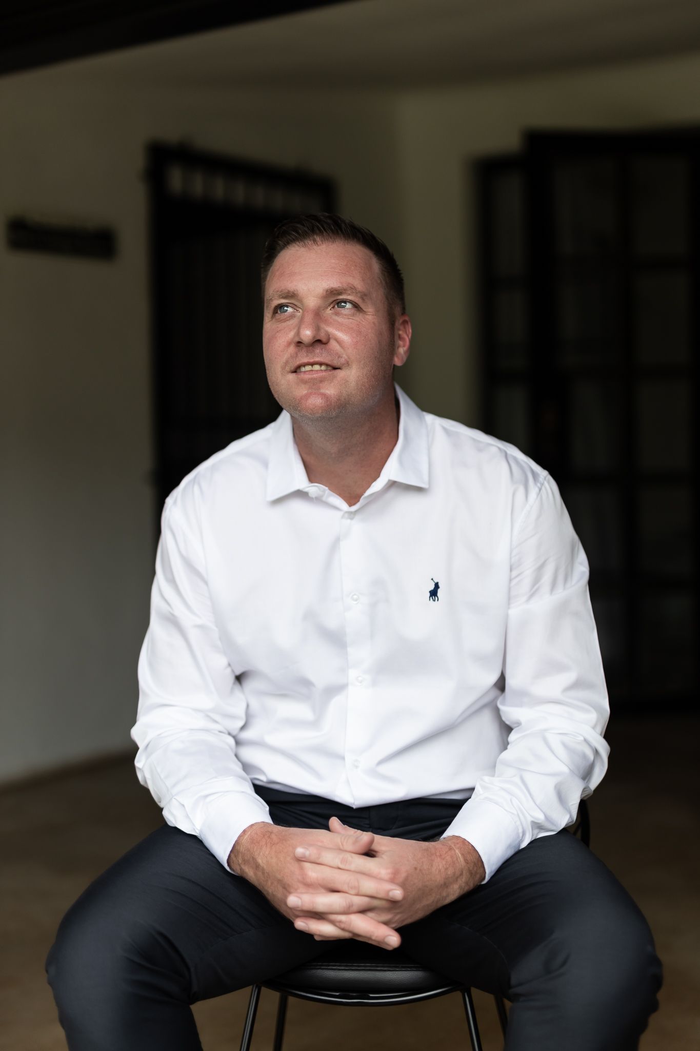 Person in white collared shirt seated on dark stool in minimal interior setting.