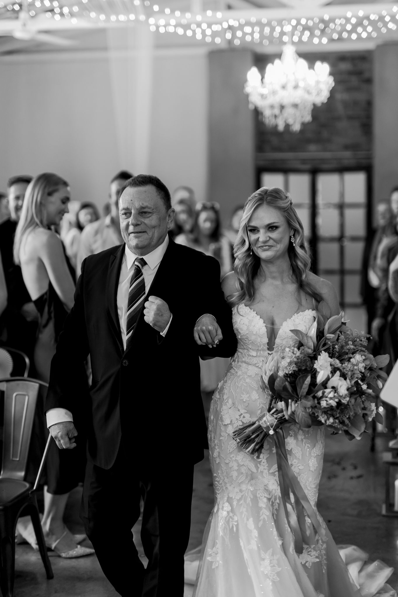 A bride in a fitted lace gown walks down the aisle with her escort at an elegant indoor ceremony.
