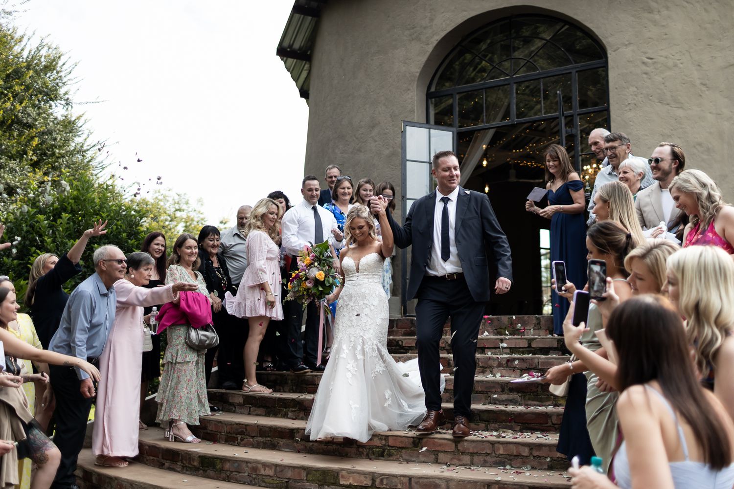 Newlyweds walk down church steps while wedding guests celebrate with confetti.