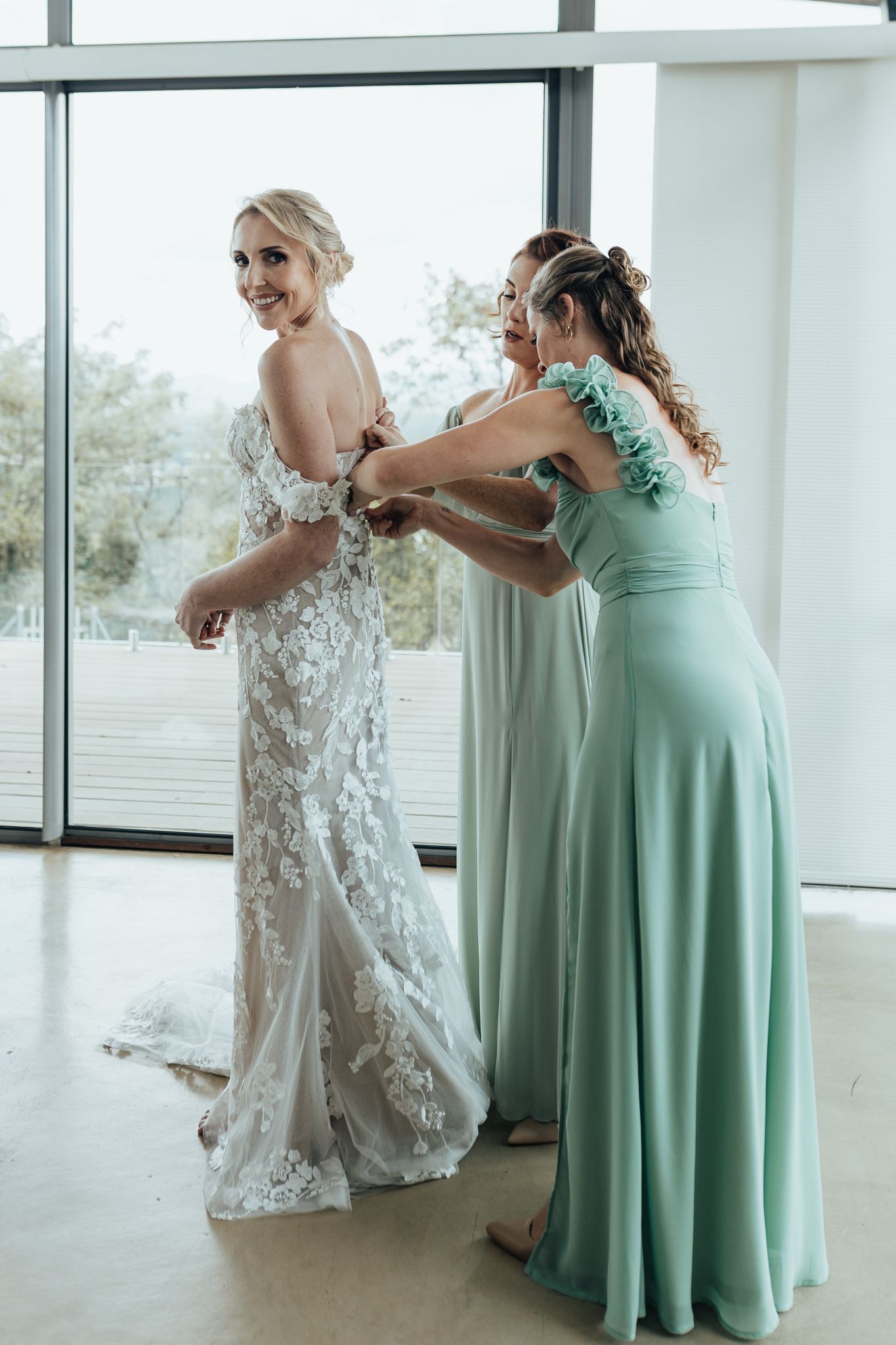 A detailed lace wedding dress being buttoned up by someone wearing a mint green bridesmaid gown in a bright room.