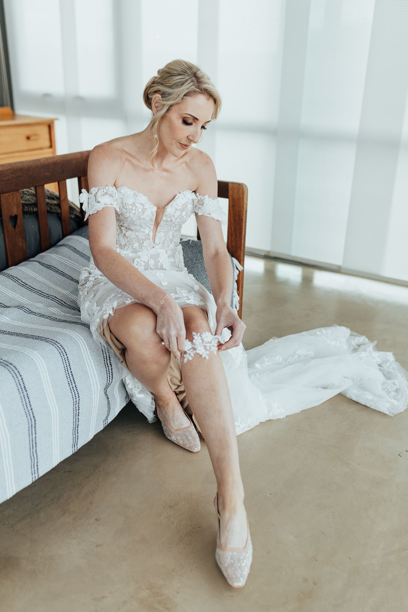 Bride in lace wedding dress sits on bed fastening her elegant white heel shoes.