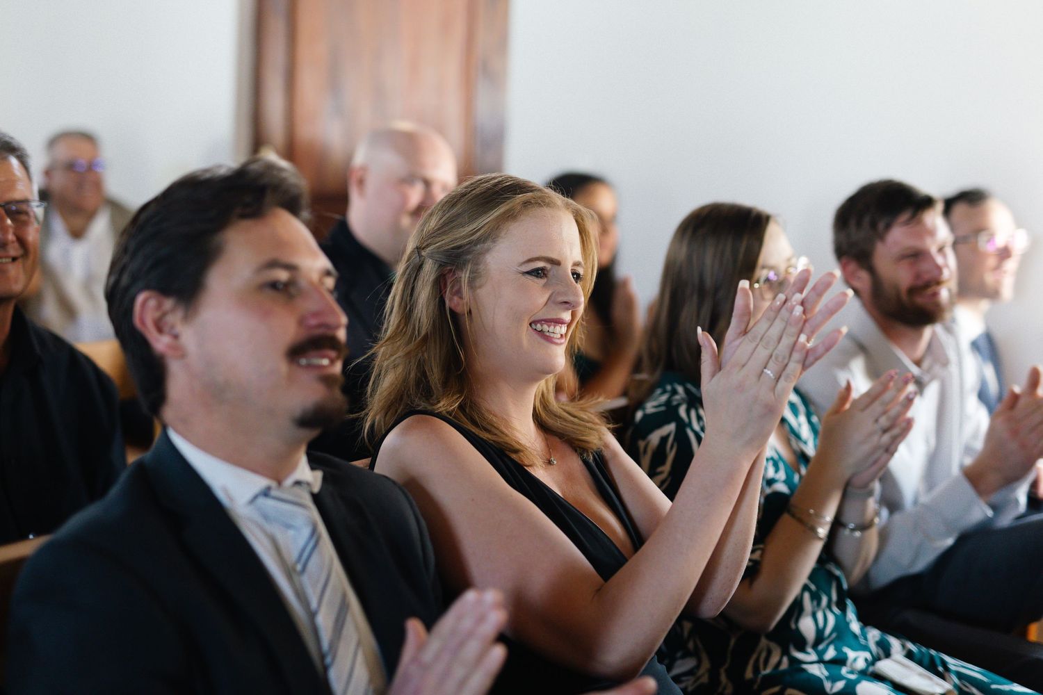 Wedding guests laugh and applaud during a joyous celebration in an indoor venue.