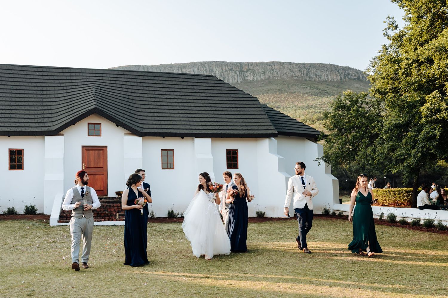 Wedding party poses in front of rustic white buildings with mountains and trees in the background.
