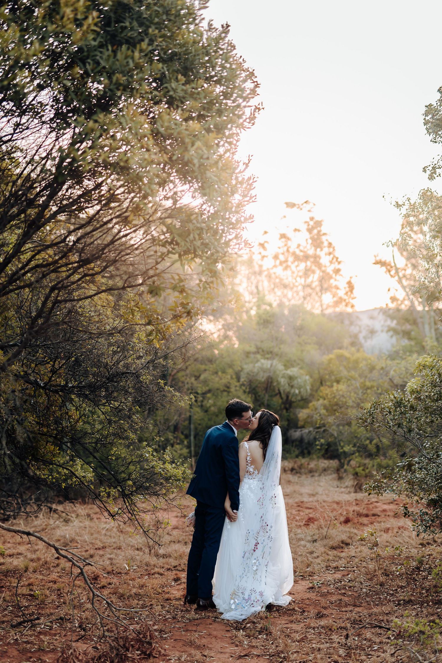 A romantic outdoor wedding scene captured at sunset with golden light filtering through trees in a desert landscape.