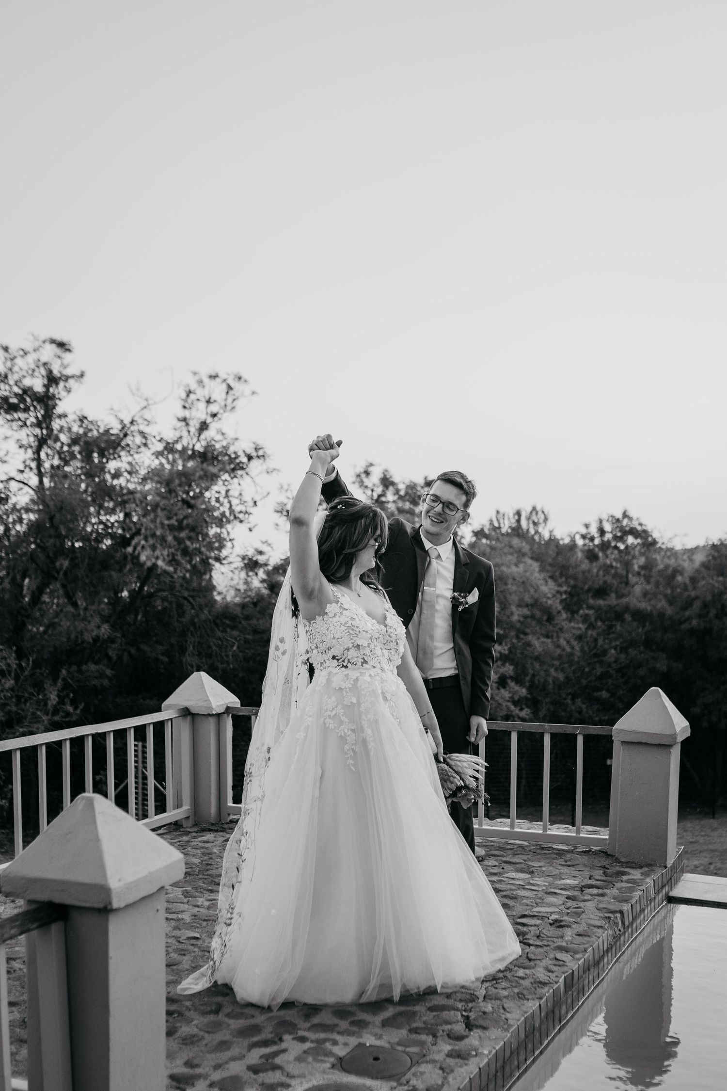 A couple in wedding attire share an intimate moment on a stone balcony surrounded by trees at sunset.