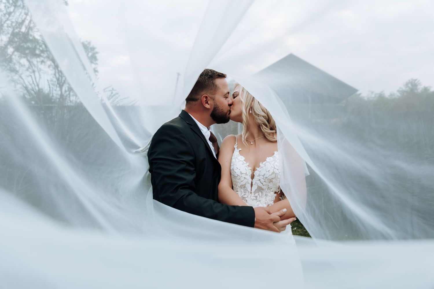 A romantic wedding couple embraces under a flowing white veil against a misty outdoor backdrop.