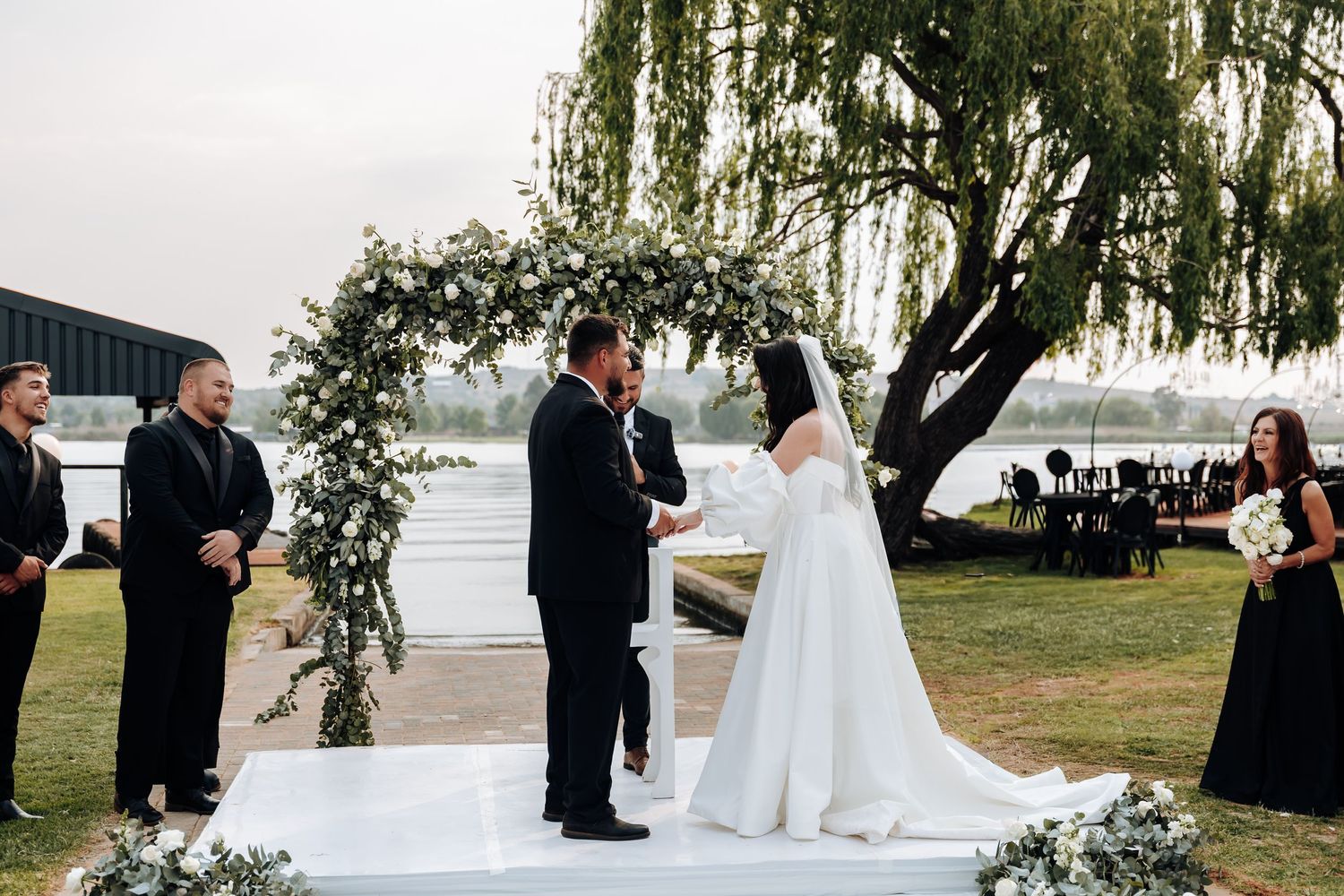 A series of outdoor wedding ceremony moments captured under a willow tree by a lake at sunset.