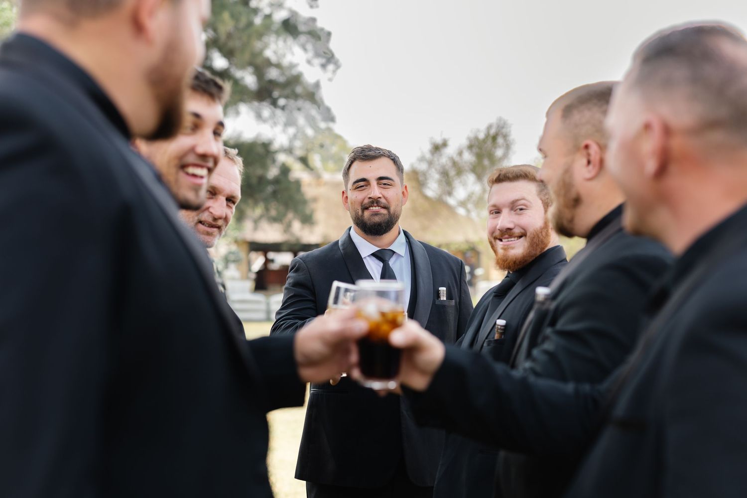 A group of groomsmen in black suits share a candid laughing moment together outdoors.