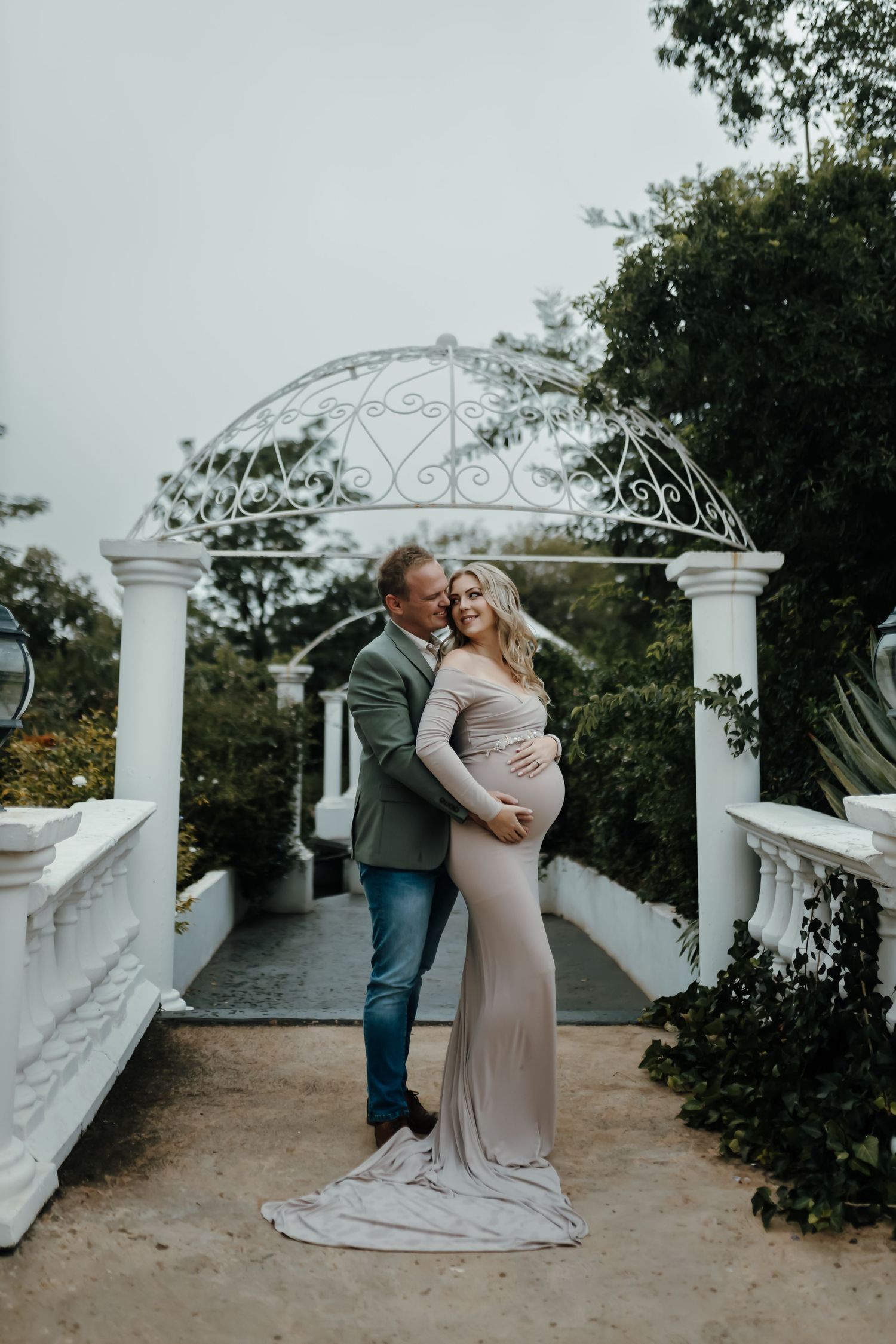 Romantic couple embracing on white terrace with decorative railings and greenery in outdoor garden setting.