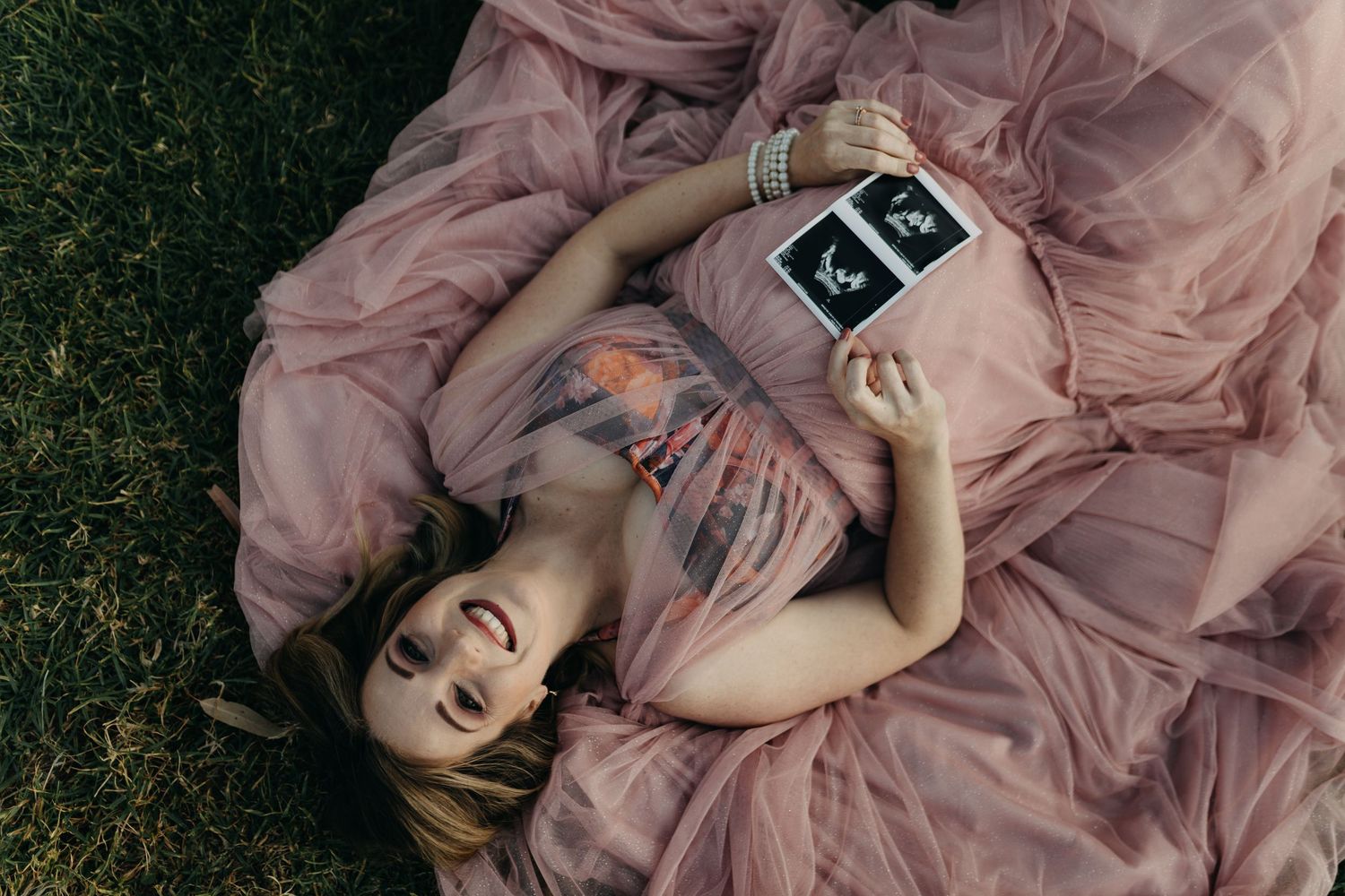 Figure in a flowing pink dress lies gracefully on grass in a romantic outdoor photoshoot setting.