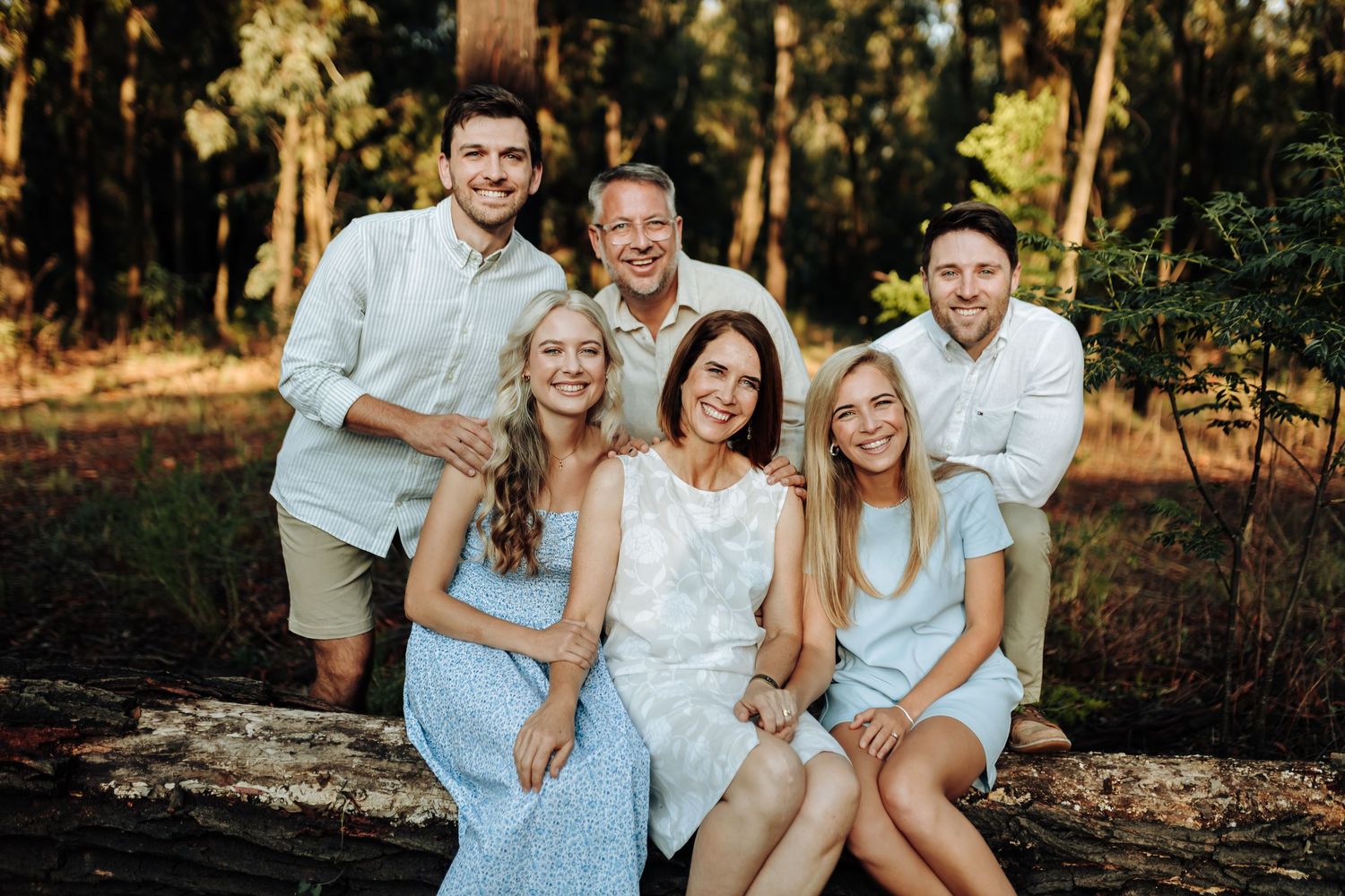 A group wearing coordinated white and blue outfits poses together in a sun-drenched wooded outdoor setting.