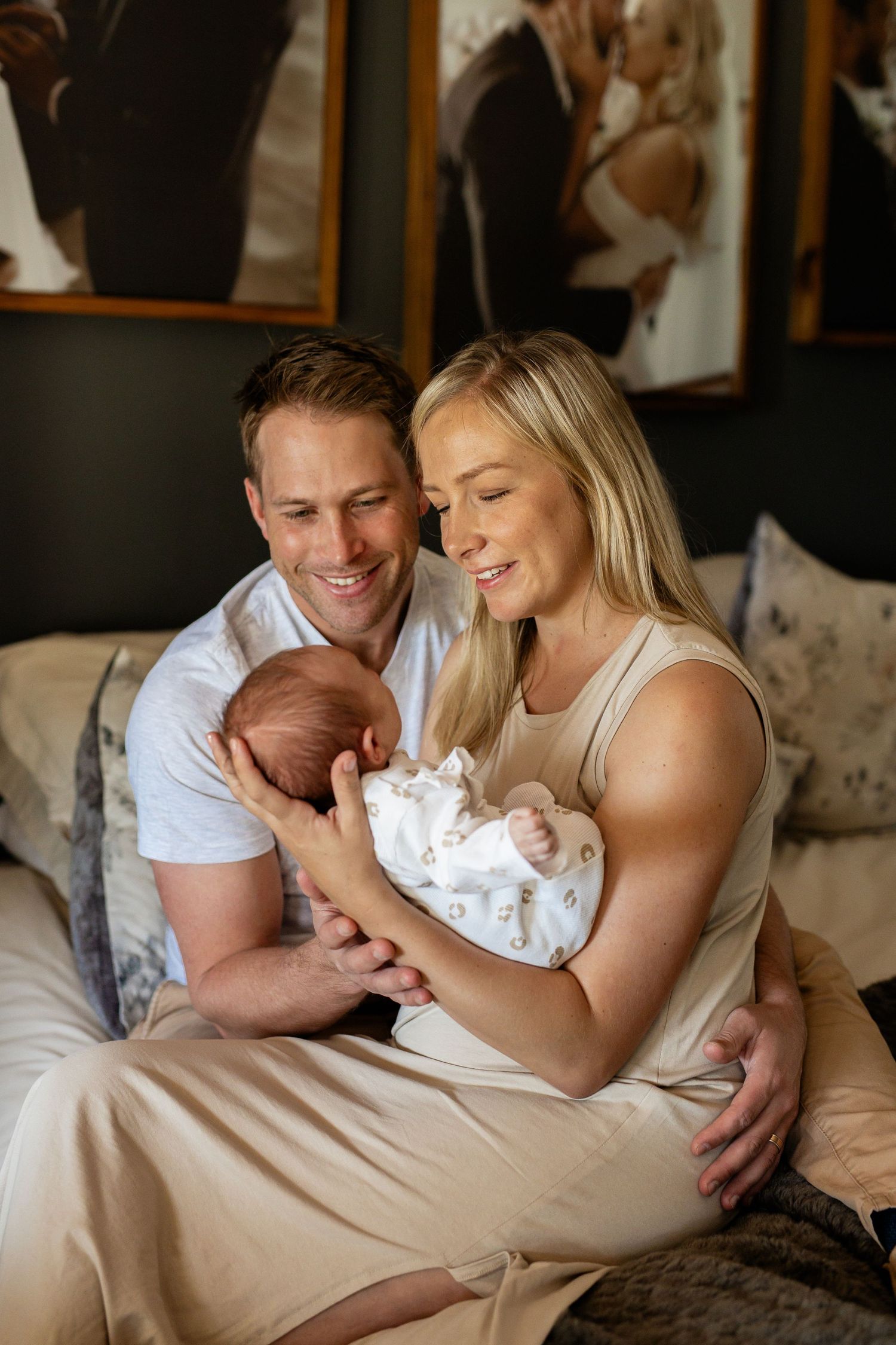 A tender family moment captured with parents snuggling their newborn baby on a bed with neutral tones and soft lighting.