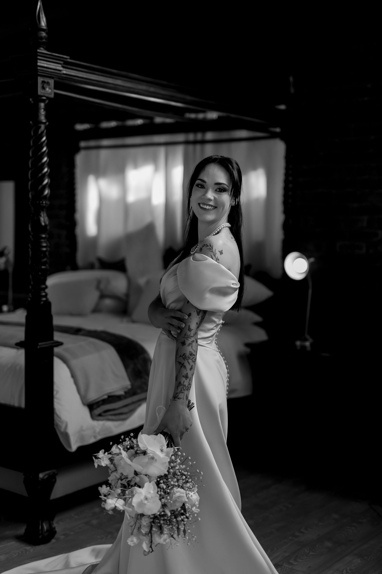 A bride in an elegant white wedding dress holds a bouquet while standing in a monochromatic bedroom setting.