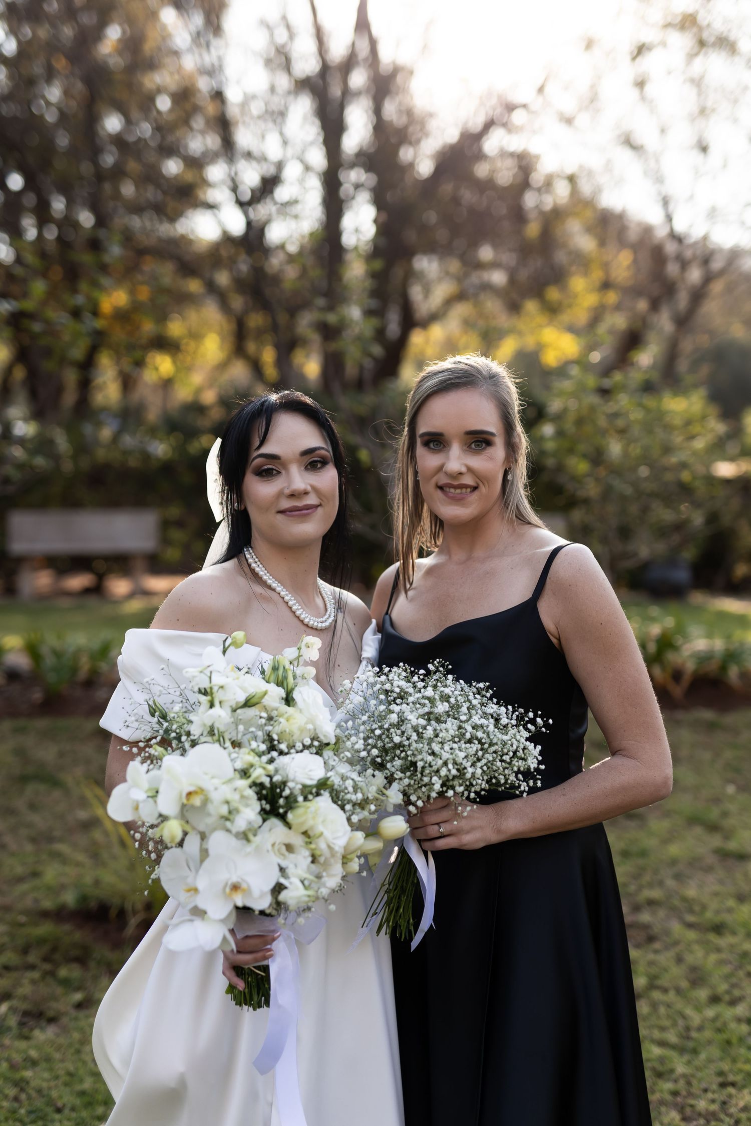 Two people pose at an outdoor wedding, one in a white wedding dress holding white orchids and one in a black formal gown.