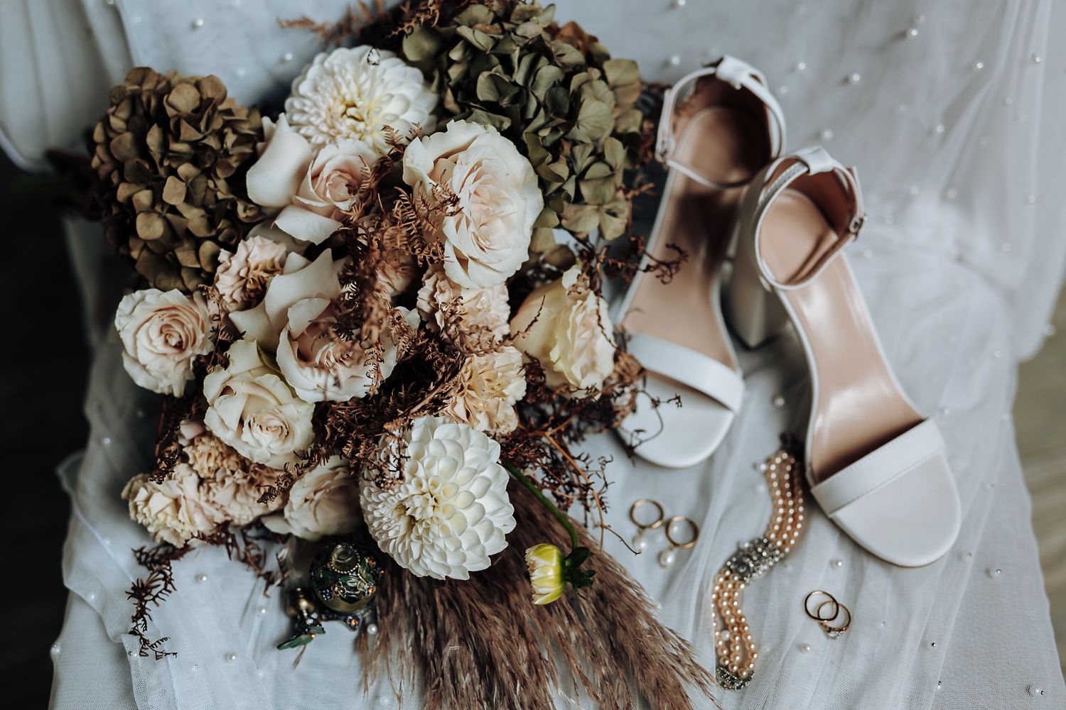 Wedding details flatlay with neutral floral bouquet, sparkly heels and delicate jewelry on white fabric backdrop.