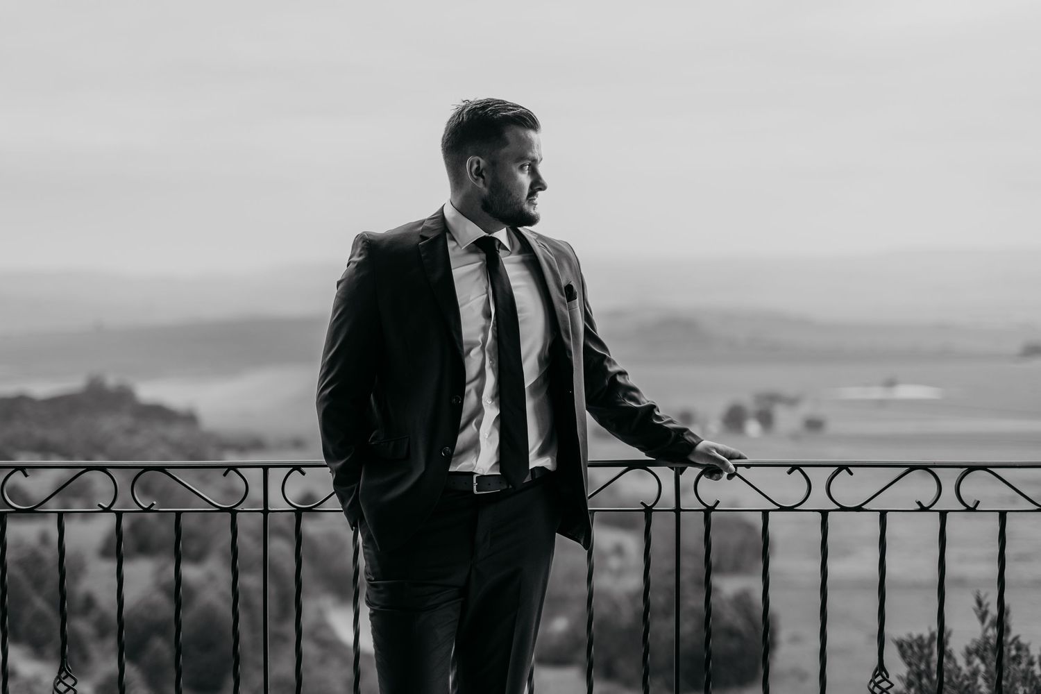 Black and white photo of a person in a suit leaning against an ornate railing with a scenic mountain view behind them.
