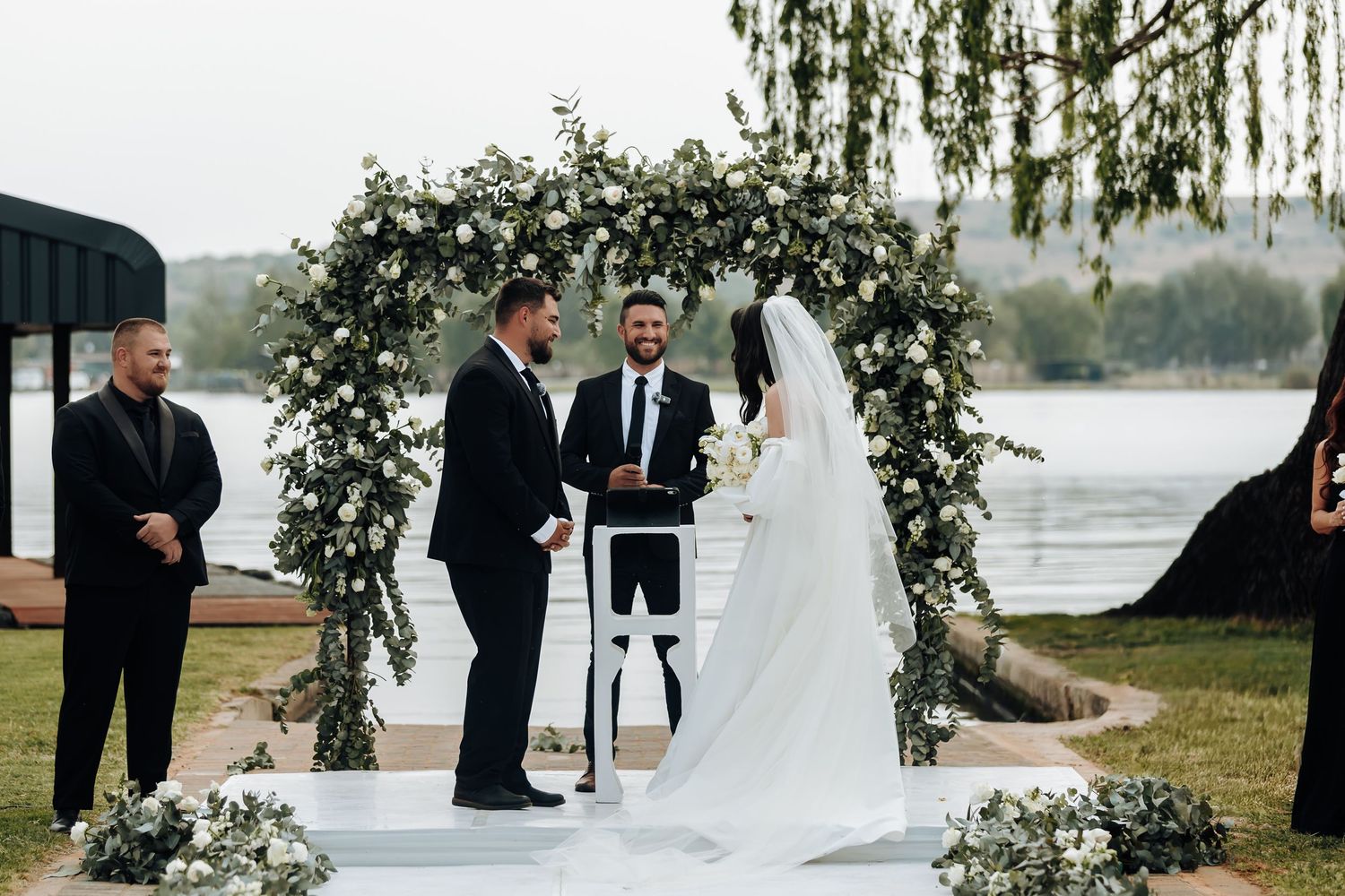 Wedding ceremony takes place under a lush greenery arch beside a tranquil lake at sunset.