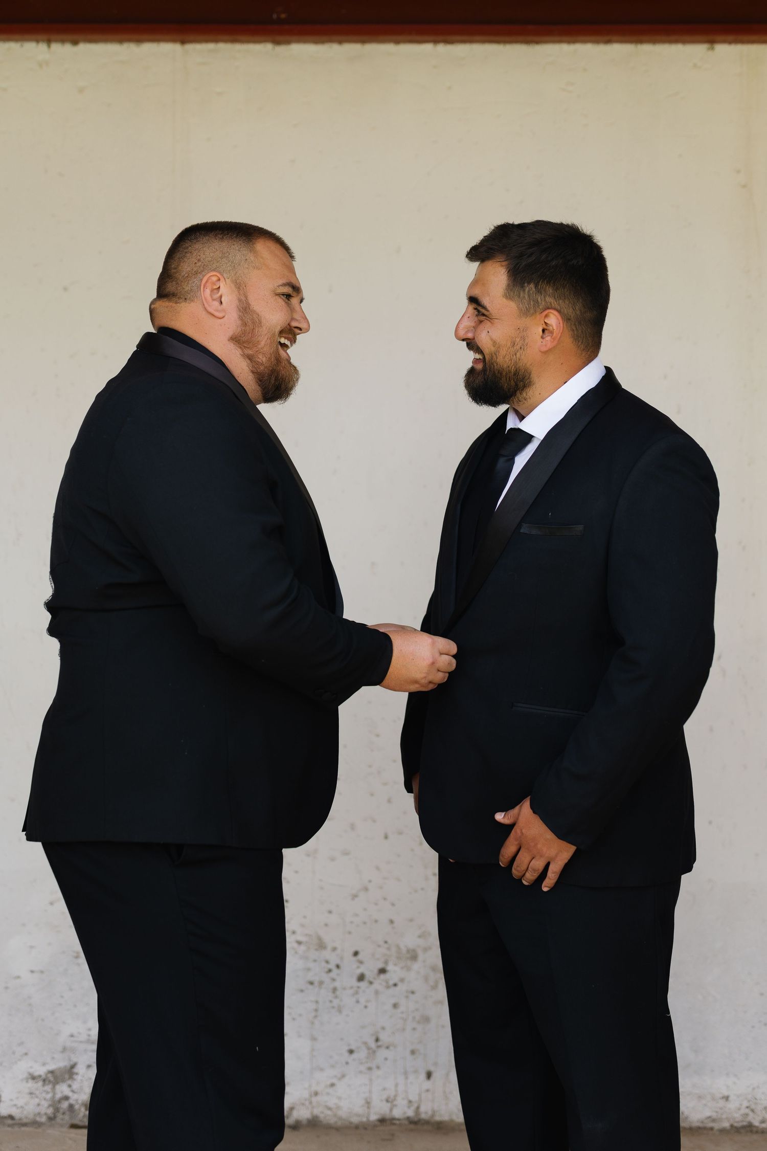 Two men in black suits share a candid moment in a series of four similar photos against a white wall.