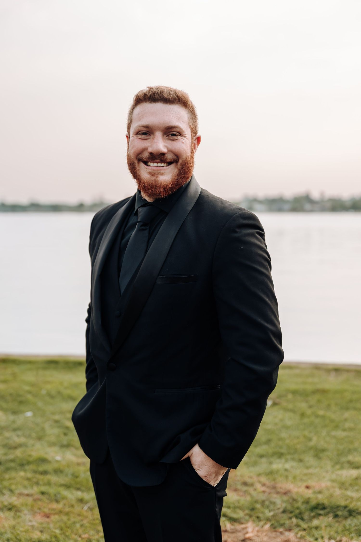 A person in a black suit stands outdoors by a waterfront on a grassy area, smiling warmly at the camera.