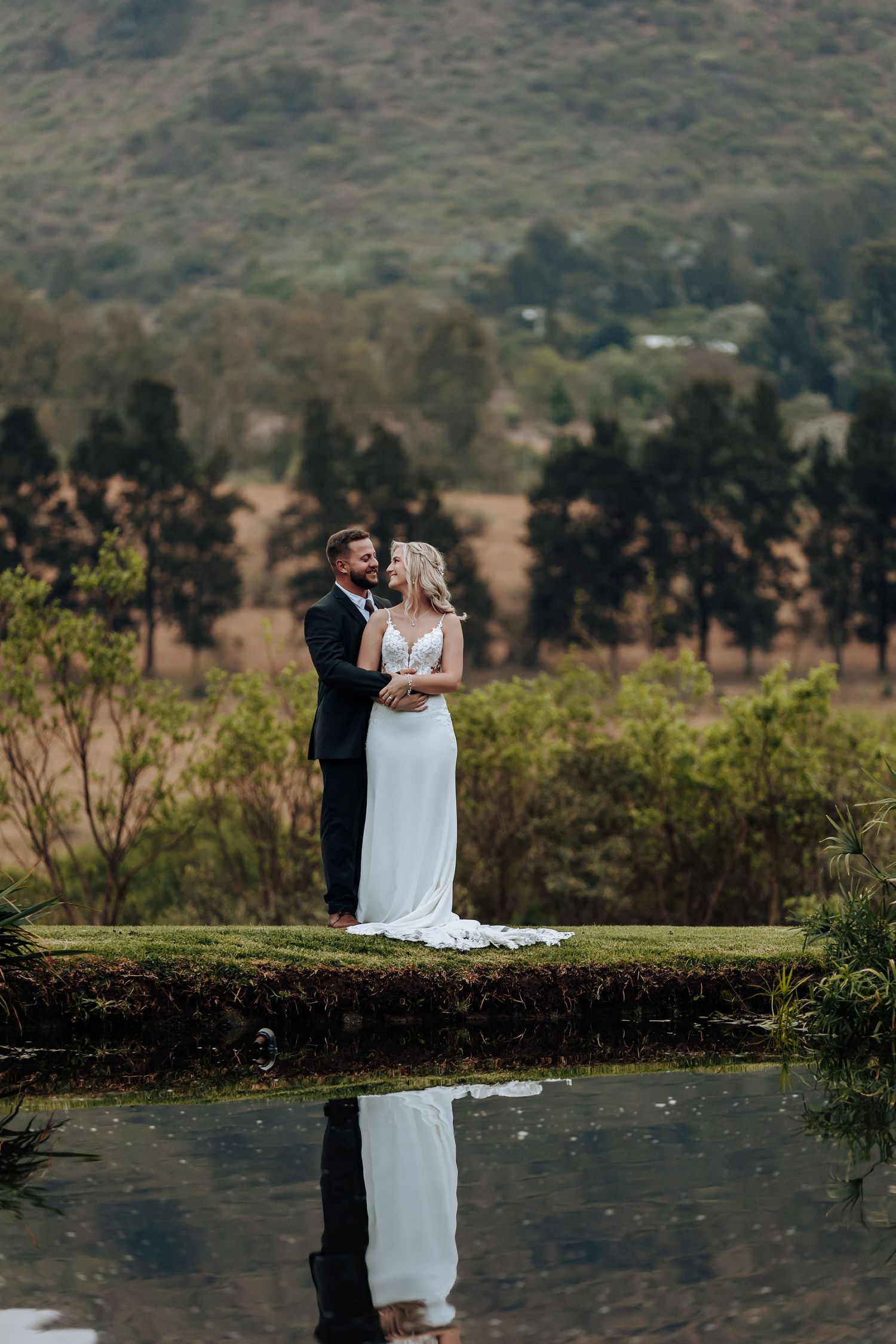 Newlywed couple embraces beside a reflective lake with mountains and trees in the background during golden hour.