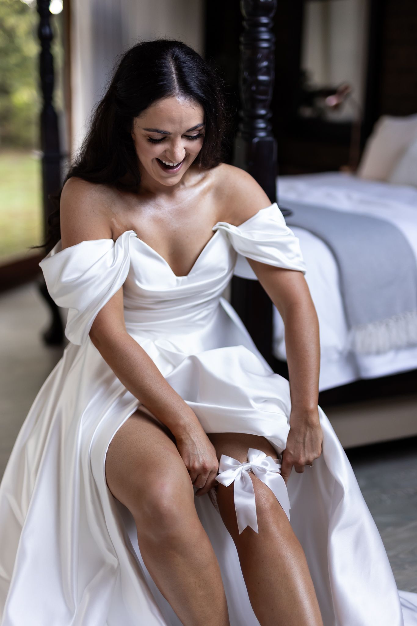 A bride in a white off-shoulder dress adjusts her garter while sitting near a canopy bed.