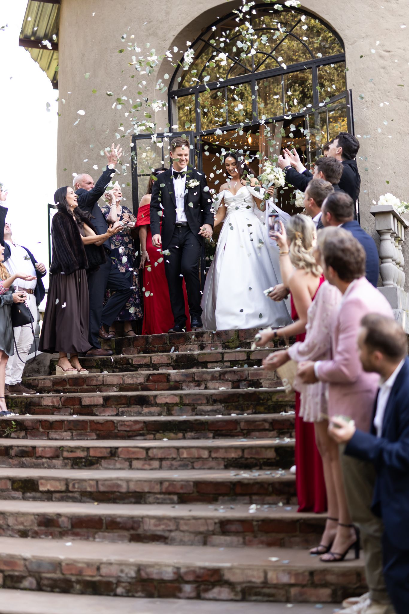 Newlyweds celebrate on brick steps as guests throw flower petals during their wedding exit.