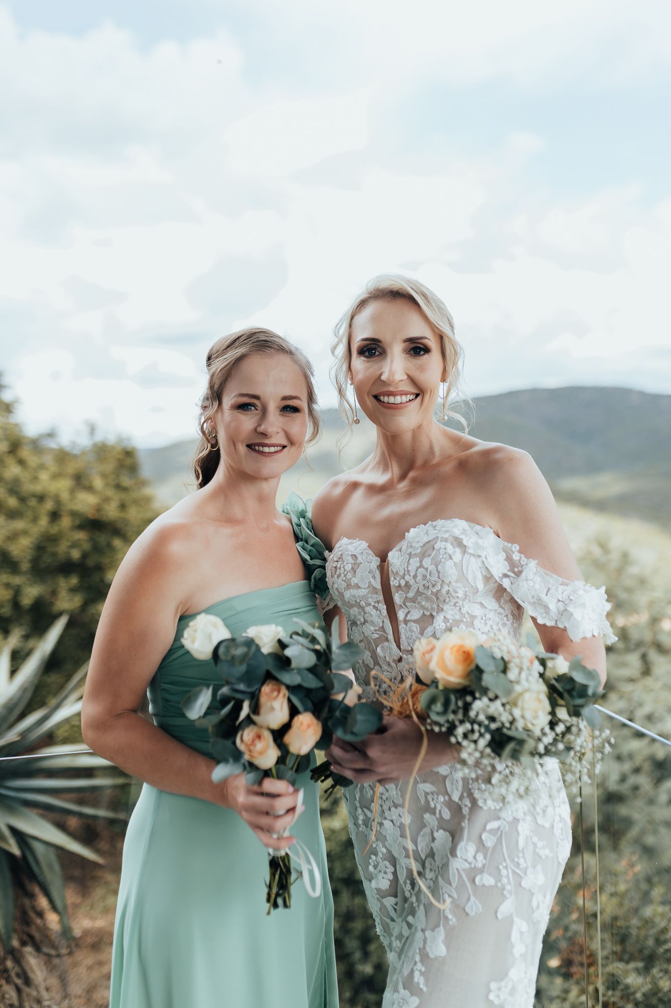 Two women in wedding attire hold floral bouquets while posing outdoors with mountain views.