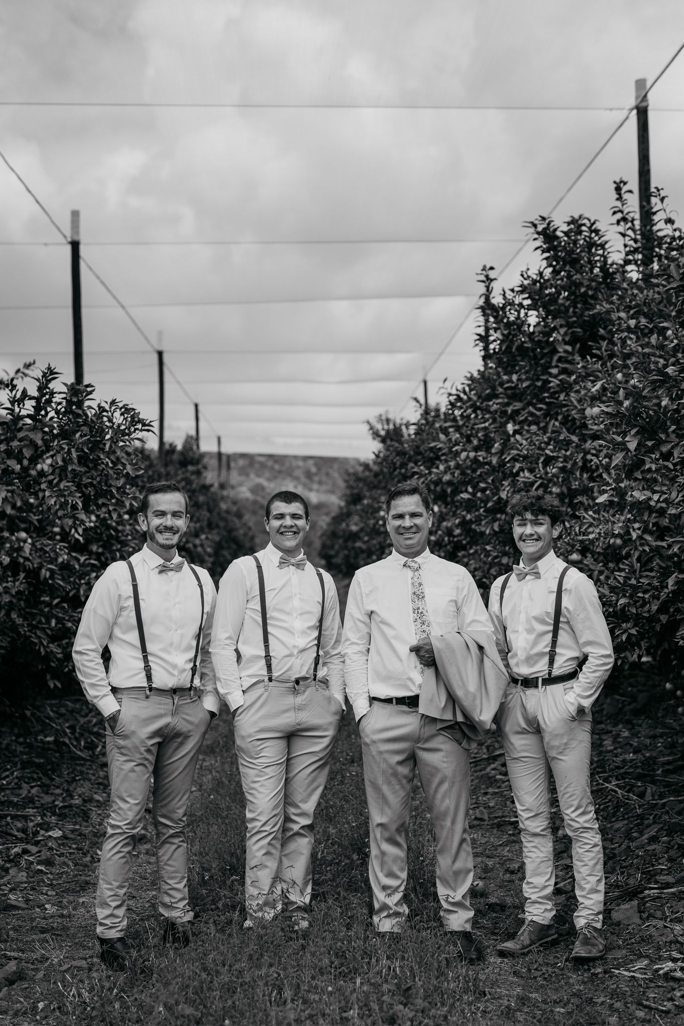 Wedding party in light colored suits with suspenders pose together in orchard path.