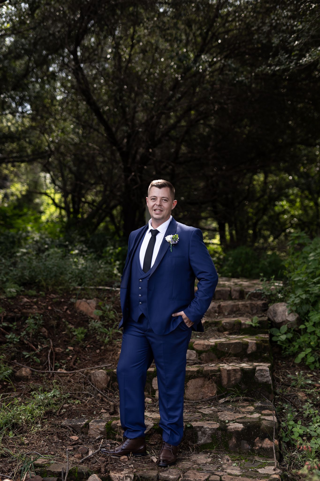 Groom in navy suit poses confidently on rustic stone steps surrounded by greenery.