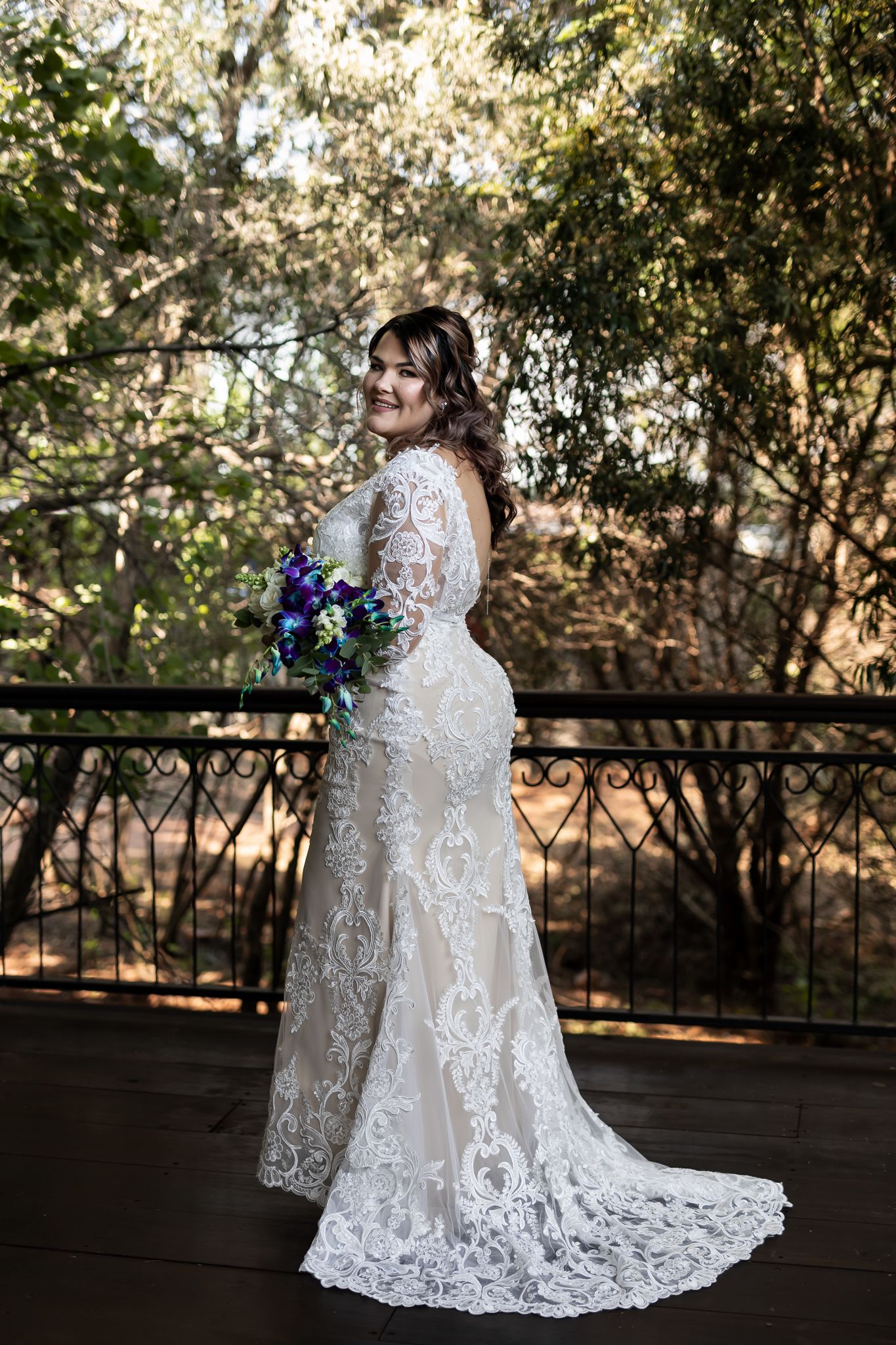 A bride in a detailed lace wedding gown poses elegantly on a wooden deck surrounded by trees.